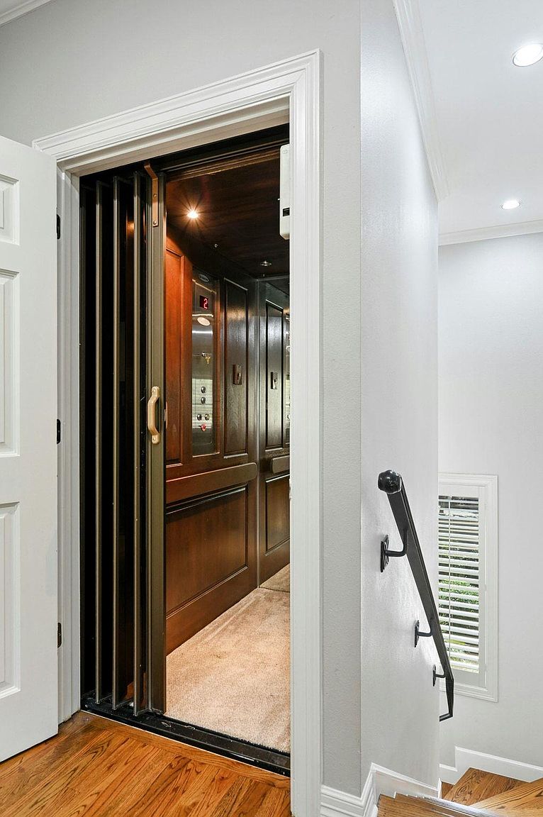 This interior shot showcases a residential elevator accessed from a hallway or stair landing. The elevator features wood paneling and a metal accordion gate, while the surrounding area includes white walls, trim, and a wooden staircase with a black handrail. The image highlights a unique accessibility feature and the home's overall design.