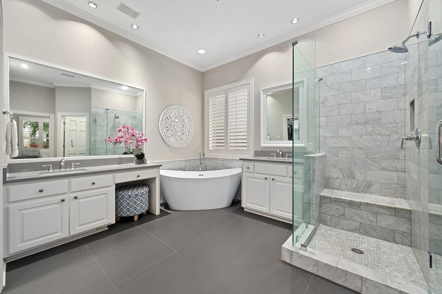 This is a luxurious primary bathroom featuring a freestanding soaking tub, a glass-enclosed shower with marble tiling, and dual vanities with white cabinetry and gray countertops. The bathroom has a neutral color palette, gray tile flooring, and ample natural light from a window with plantation shutters. The overall impression is clean, modern, and spa-like.