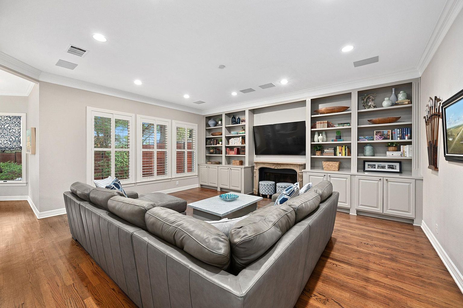 This is an interior shot of a living room featuring a large, gray sectional sofa facing a built-in entertainment center with a television and shelving units. The room has hardwood floors, recessed lighting, and windows with shutters, creating a bright and inviting atmosphere. The overall style is traditional with a touch of modern comfort.