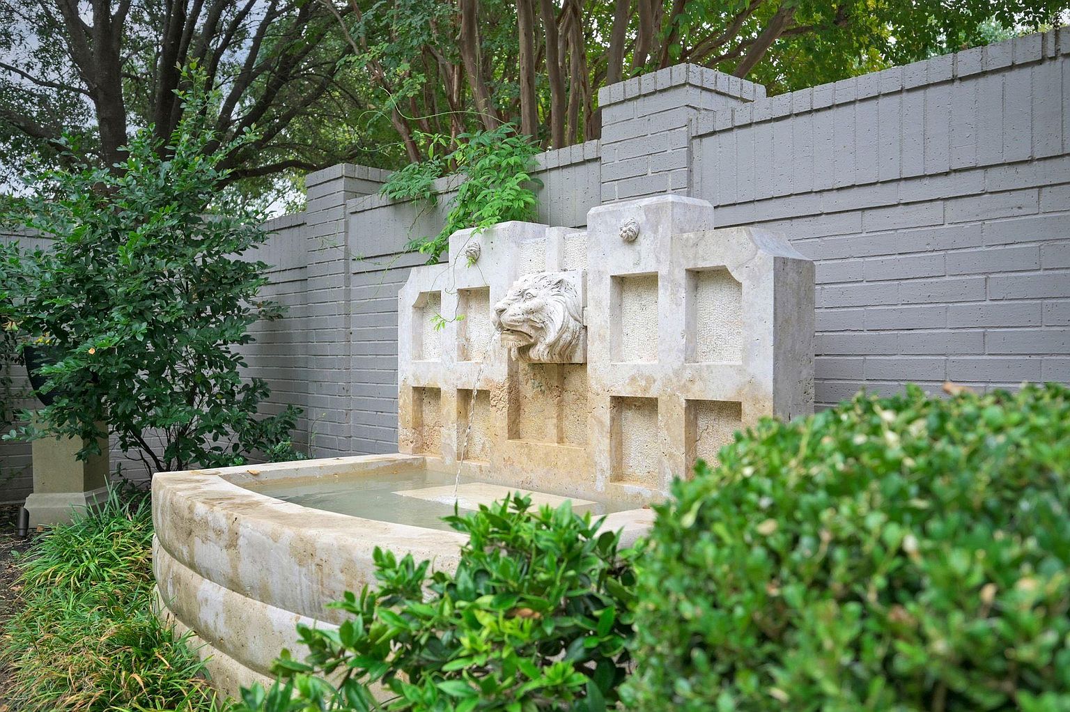 This image showcases a beautifully landscaped yard featuring a stone fountain with a lion's head spout. The fountain is surrounded by lush greenery, including bushes and trees, creating a serene and inviting atmosphere. A brick wall serves as a backdrop, adding texture and privacy to the outdoor space.
