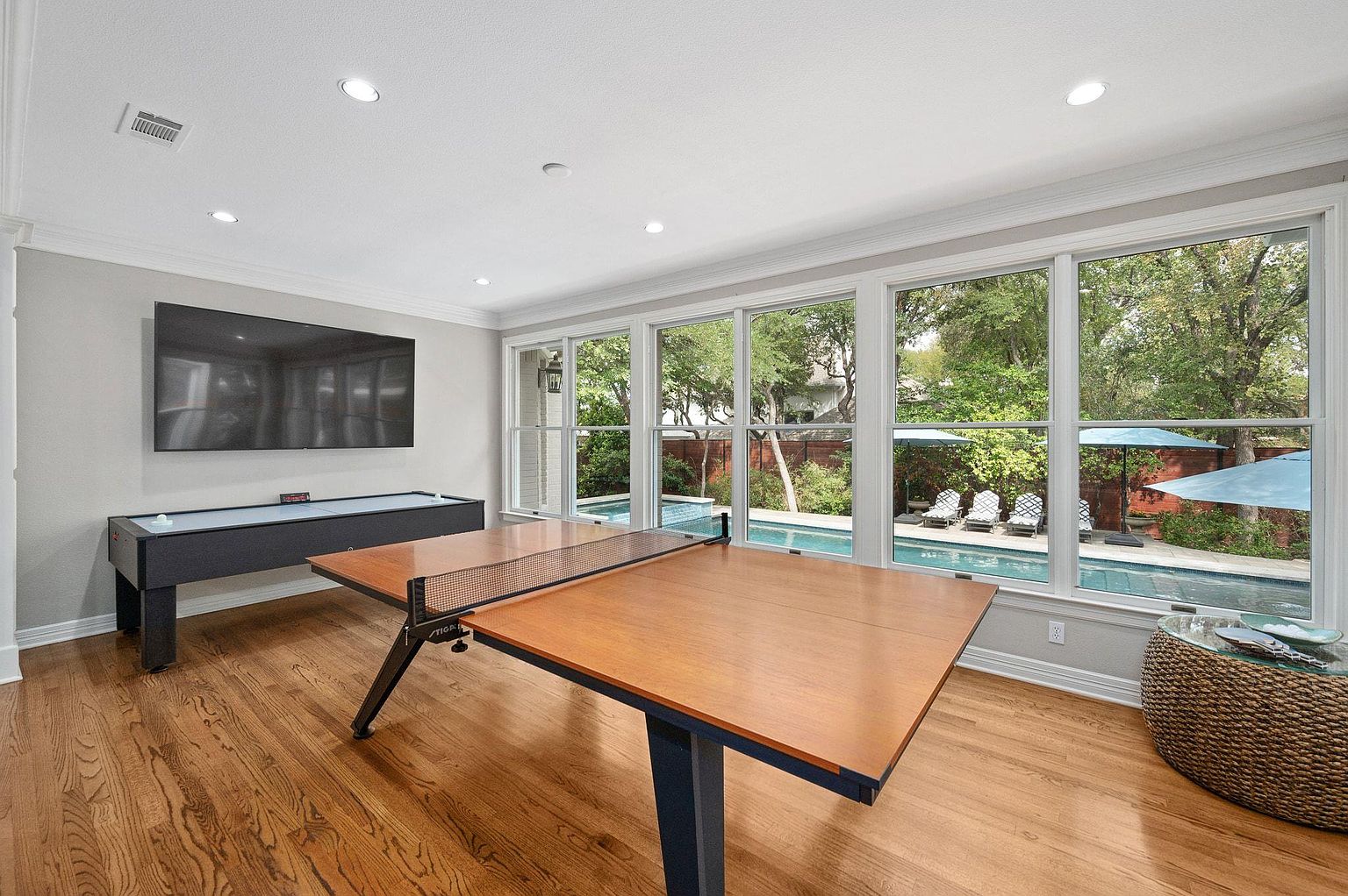 This sunroom features a ping pong table and an air hockey table, suggesting a recreational space. Large windows offer a view of the outdoor pool and patio area, bringing natural light into the room. The hardwood floors and neutral wall colors create a bright and inviting atmosphere.