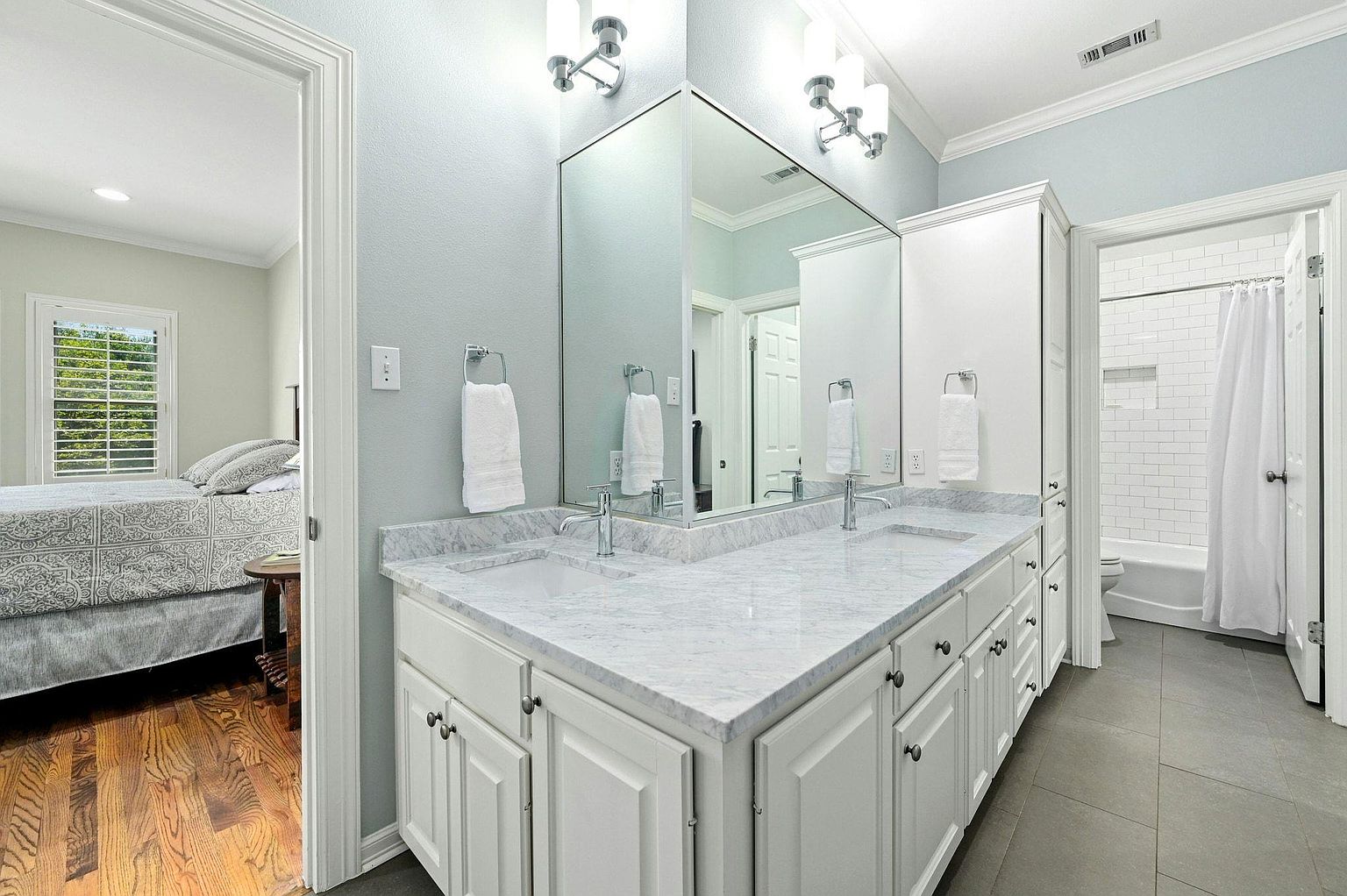 This is a well-lit primary bathroom featuring a double vanity with a white marble countertop and white cabinets. A large mirror spans the length of the vanity, reflecting the light and creating a spacious feel. The bathroom also includes a separate toilet and shower area, enhancing privacy and functionality.