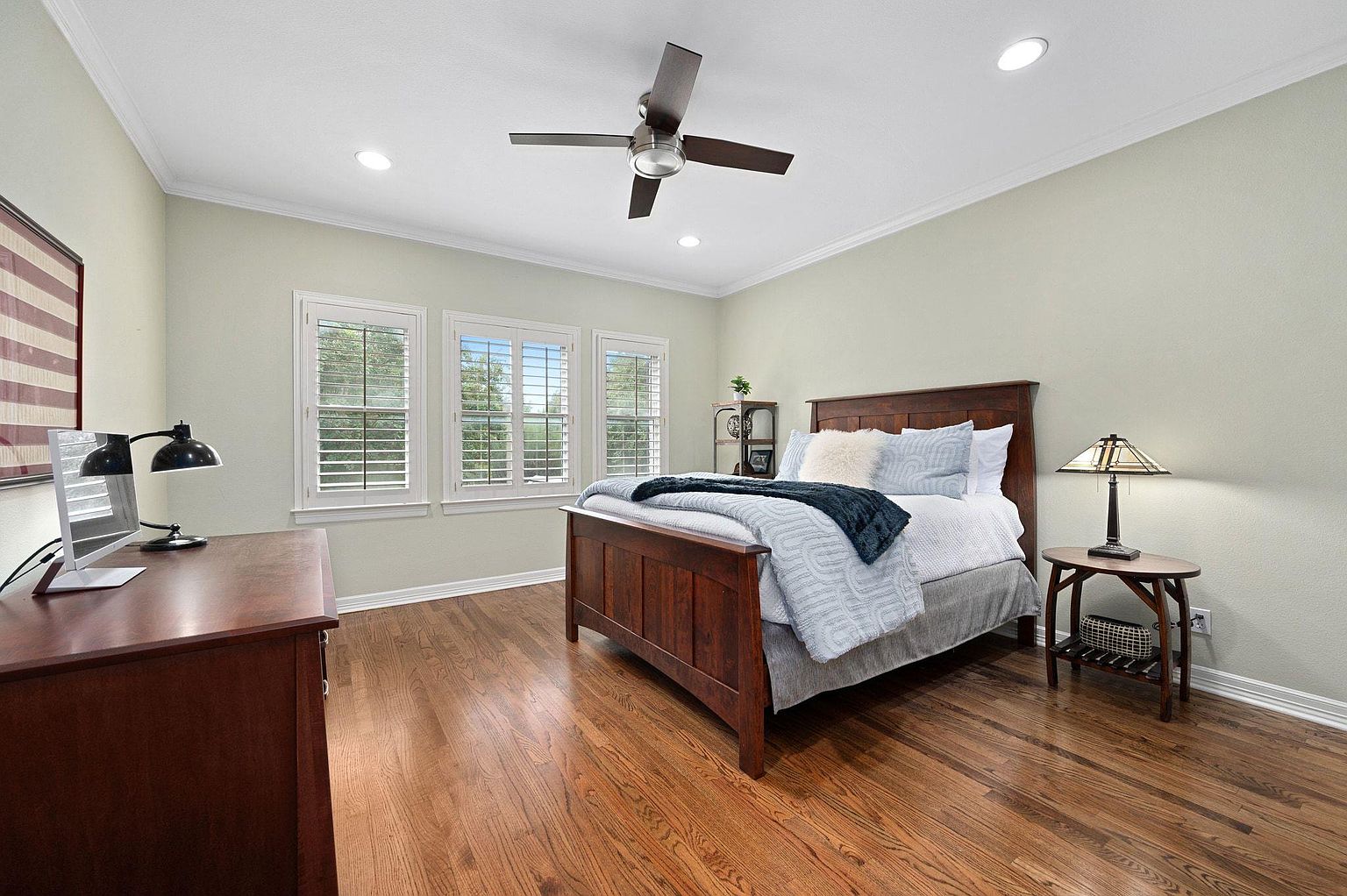 This is a well-lit primary bedroom featuring a wooden bed frame with blue and white bedding, complemented by a dark blue throw. The room has hardwood floors, light green walls, and three windows with white shutters, providing ample natural light. A dark wood dresser and a small side table with a lamp add to the room's functionality and charm.