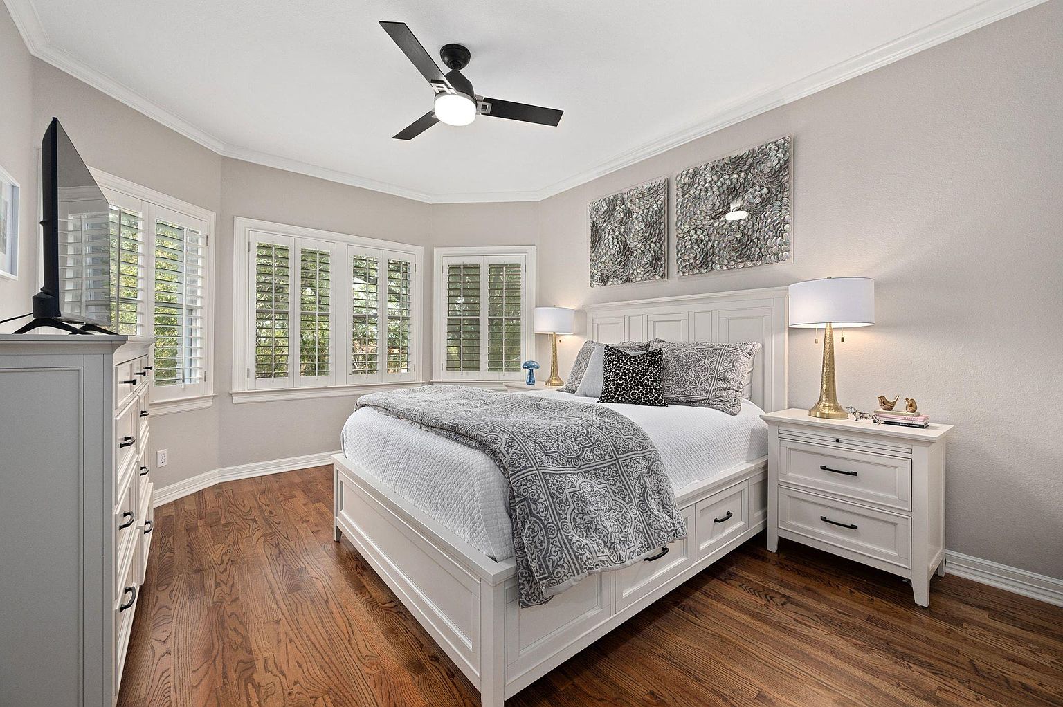 This is a well-lit primary bedroom featuring a white bed with a patterned gray throw, decorative pillows, and matching white nightstands. The room has hardwood floors, neutral-colored walls, and two pieces of artwork above the bed. Natural light streams in through the windows with white shutters, creating a bright and inviting space.
