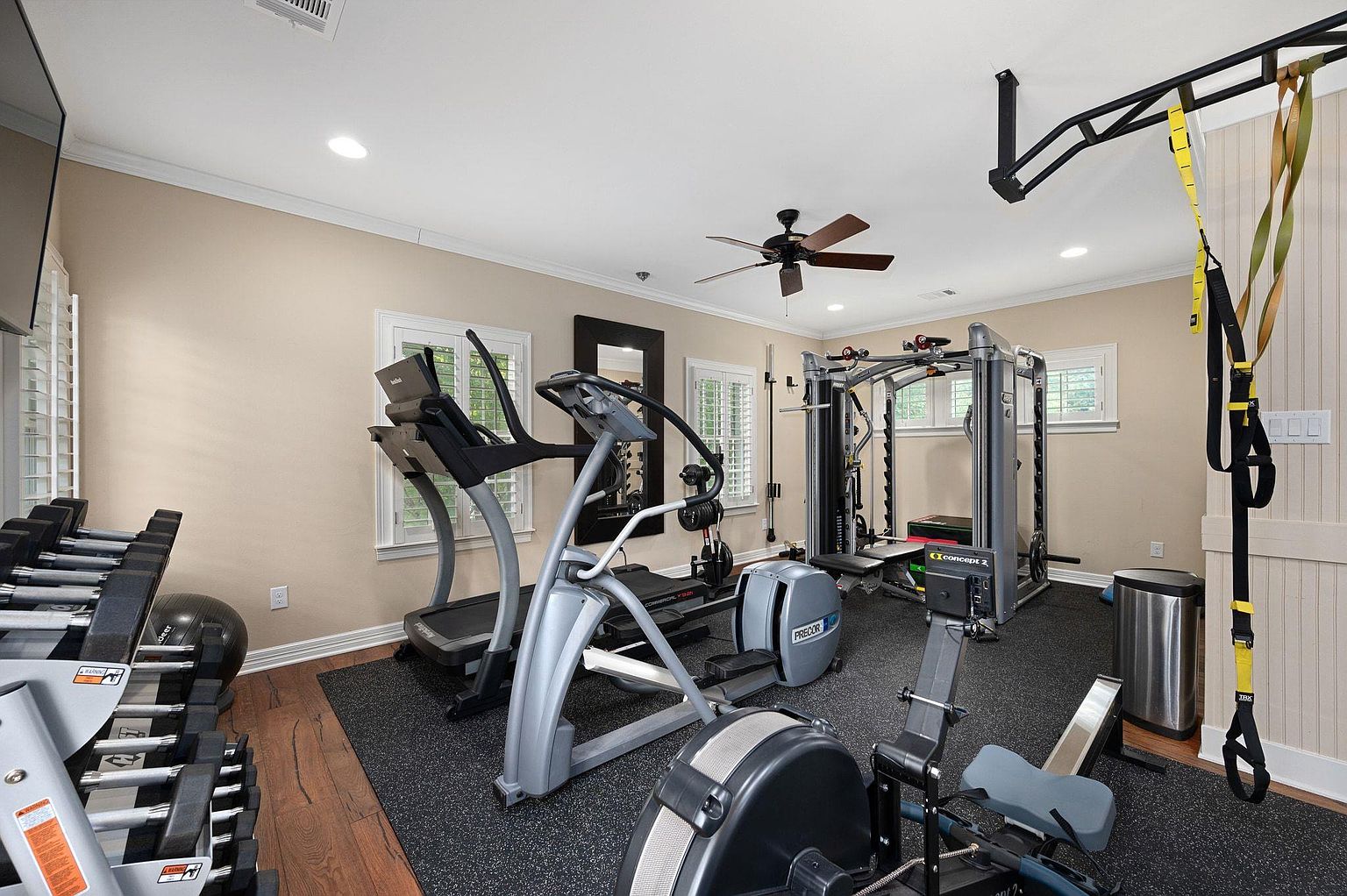 This is an interior shot of a well-equipped home gym. The room features a variety of exercise equipment including a treadmill, elliptical, rowing machine, and a weight rack with dumbbells. The flooring is a dark rubber material, and the walls are painted a neutral beige, creating a functional and inviting workout space.