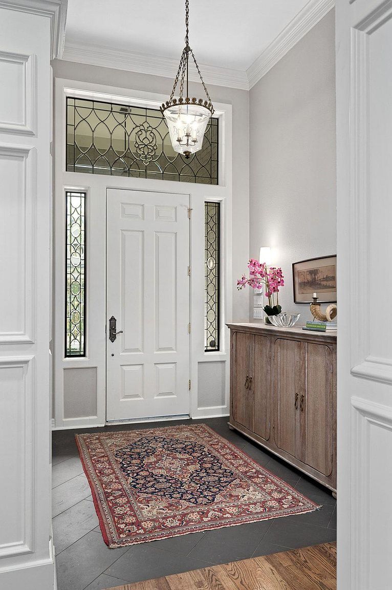 This is an interior shot of a home's entryway, featuring a white front door flanked by decorative sidelights and topped with a transom window. A patterned rug lies on the dark tile floor, leading into the home. A wooden cabinet sits to the right, adorned with a lamp, flowers, and decorative objects, adding a touch of elegance to the space.