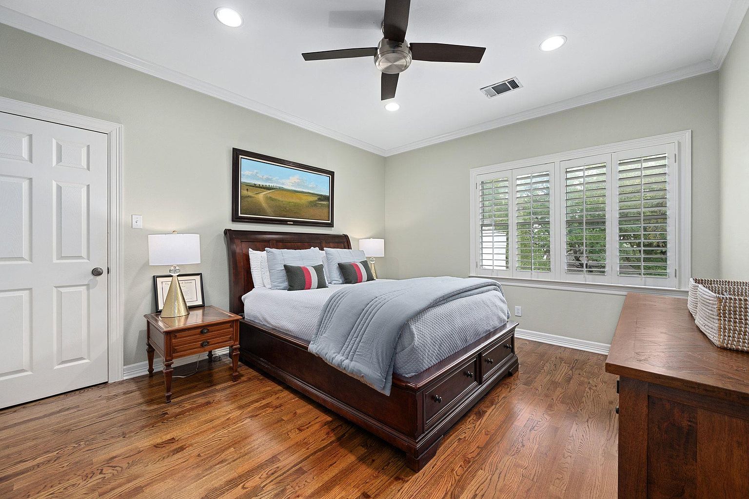 This is a primary bedroom featuring a dark wood bed frame with storage drawers, complemented by hardwood flooring. The room is well-lit with natural light from a window with white shutters and recessed lighting. A landscape painting hangs above the bed, adding a touch of color to the neutral walls.
