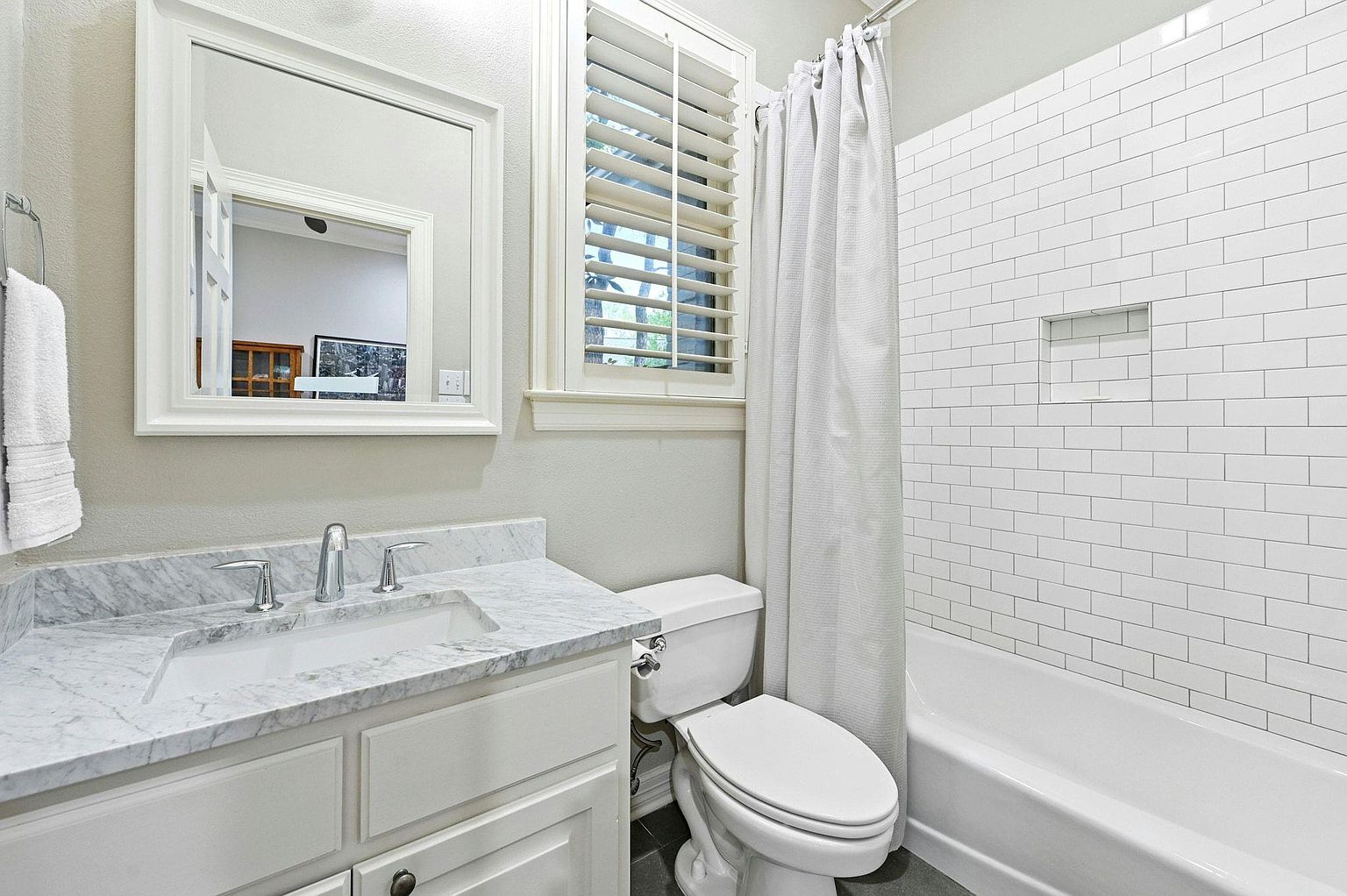 This is a well-lit bathroom featuring a white vanity with a marble countertop, a rectangular mirror, and a window with white shutters. The bathtub and shower area are tiled in white subway tiles, complemented by a neutral-colored shower curtain. The toilet is also white, and the overall impression is clean and bright.