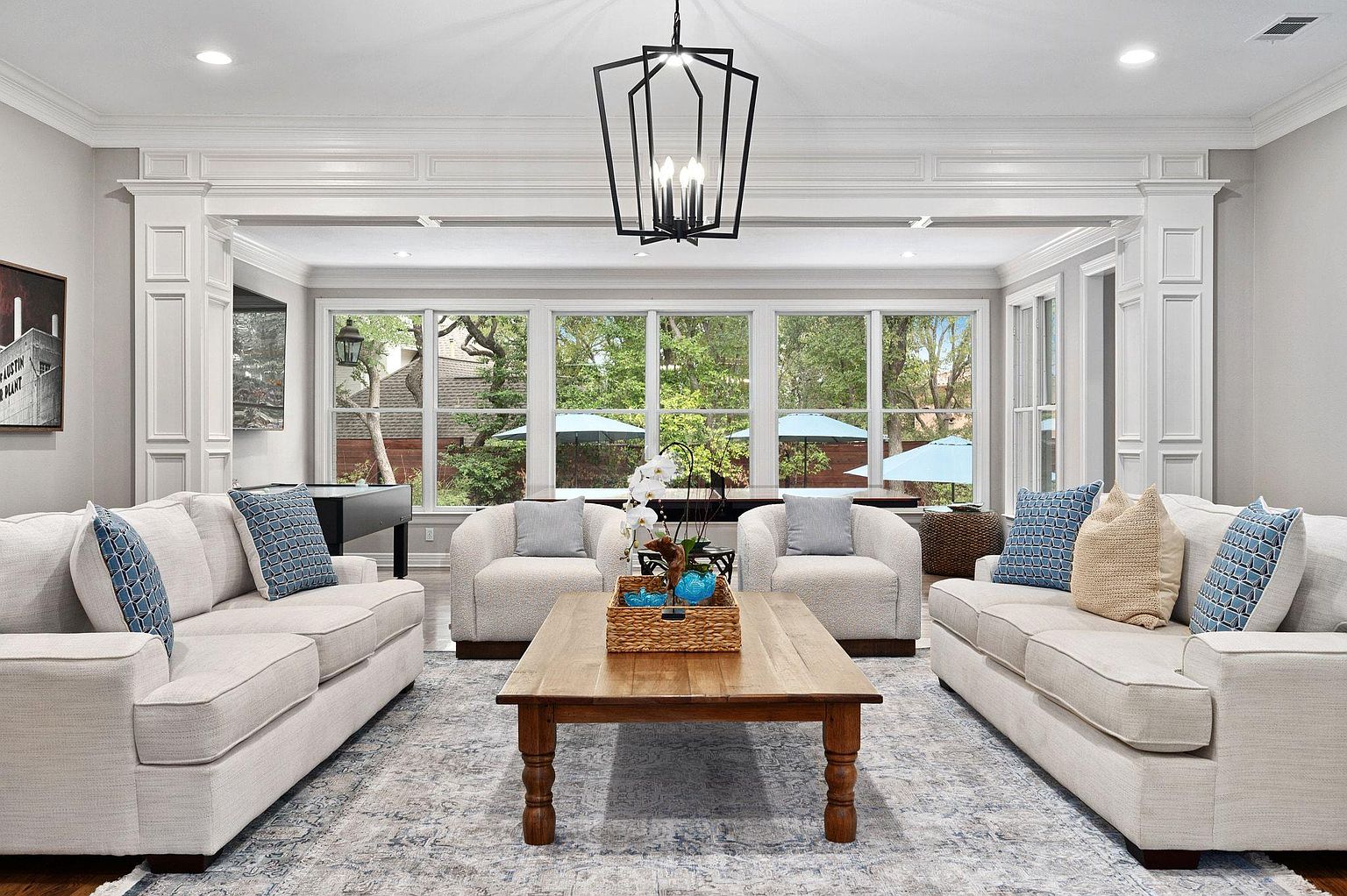 This is a well-lit living room featuring two light-colored sofas with patterned throw pillows, two armchairs, and a large wooden coffee table. A modern black chandelier hangs from the ceiling, and large windows offer a view of the outdoors. The room is spacious and elegantly decorated, creating a comfortable and inviting atmosphere.