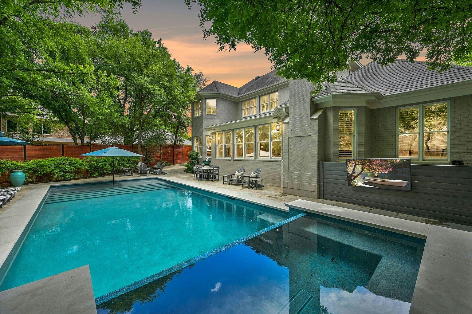 This image showcases a luxurious backyard pool and spa area. The pool features clear blue water and is surrounded by a patio with lounge chairs and an umbrella. The house, visible in the background, has large windows and a well-maintained exterior, suggesting a high-end property perfect for relaxation and entertainment.