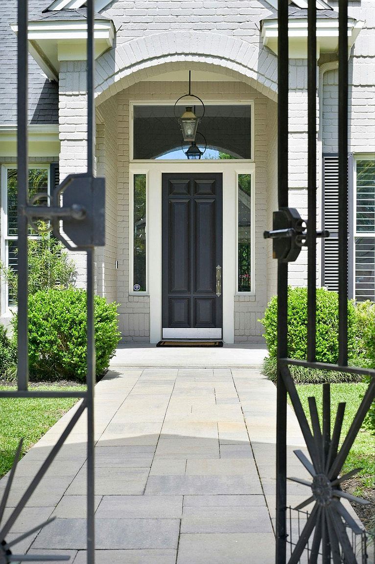 This image showcases the entryway of a home, viewed through an ornate iron gate. The pathway leads to a dark-colored front door framed by sidelights and a transom window, with a hanging lantern above. Lush greenery flanks the walkway, creating a welcoming and well-maintained appearance.