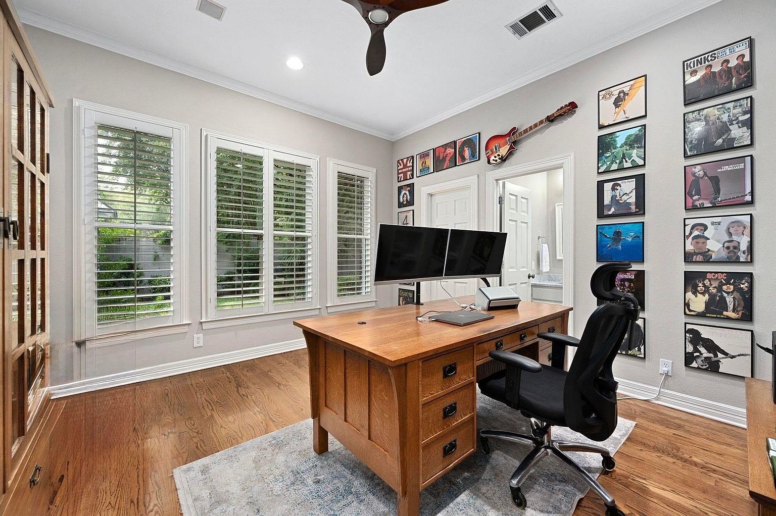This is an interior shot of a home office featuring a large wooden desk with dual monitors, a black office chair, and a wall adorned with framed album covers and a guitar. Natural light floods the room through shuttered windows, highlighting the hardwood floors and a patterned area rug. The space appears organized and conducive to productivity, with a touch of personal style.