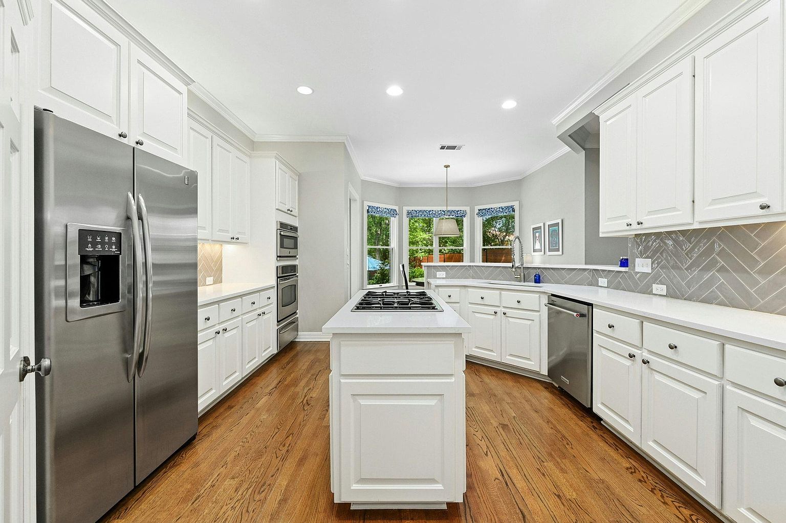 This is a bright and spacious kitchen featuring white cabinetry, stainless steel appliances, and hardwood floors. A central island with a cooktop provides ample workspace, while a bay window offers natural light and a view of the outdoors. The kitchen's design is both functional and aesthetically pleasing, creating a welcoming atmosphere.