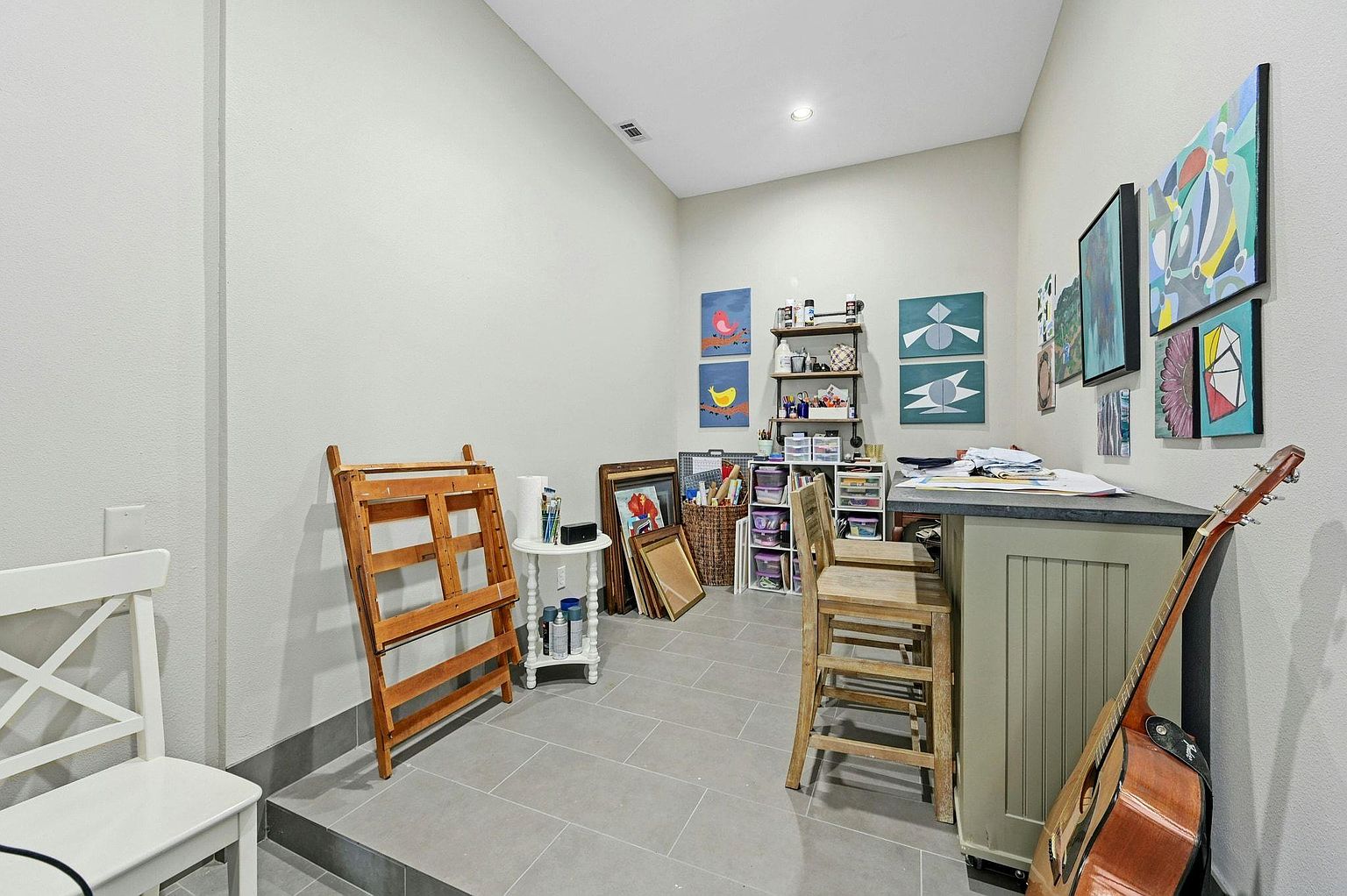 This is an interior shot of a home office or study. The room features a neutral color palette with gray tile flooring and light gray walls. There is a desk area with bar stools, artwork displayed on the walls, and shelving units filled with various items, suggesting a creative or hobby space.