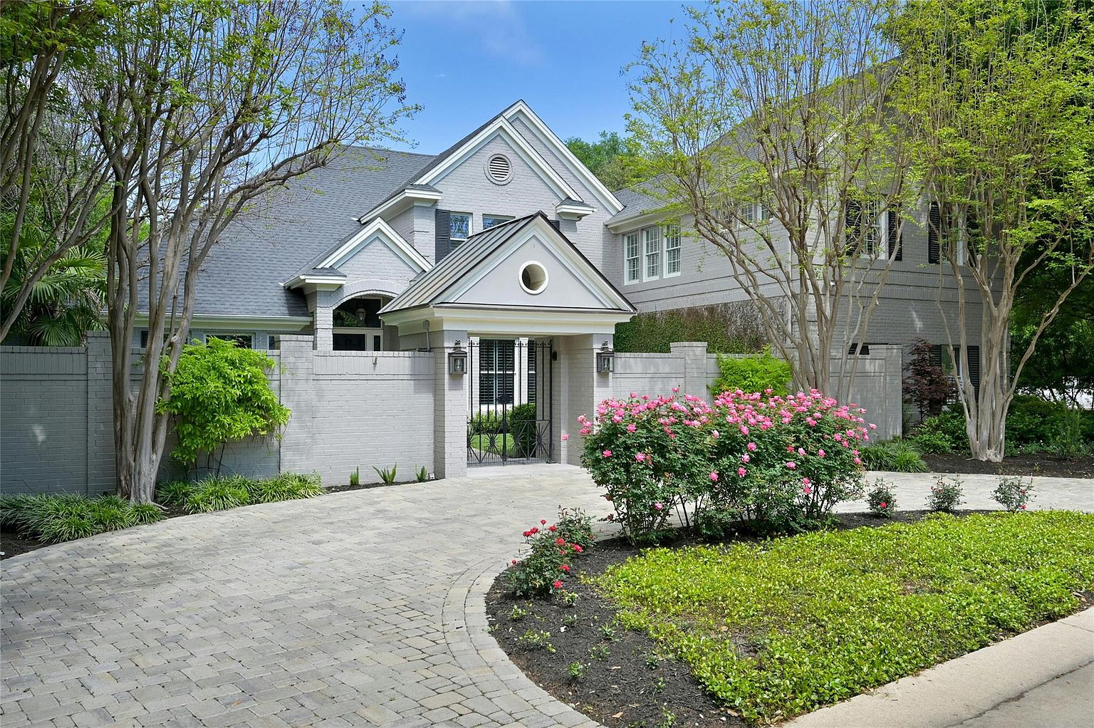 This is a front view of a well-maintained, single-family home featuring a gray brick facade, a dark gray roof, and a charming gated entryway. The property boasts mature trees and manicured landscaping, including a vibrant rose bush and neatly trimmed greenery. The circular driveway is paved with interlocking stones, adding to the home's curb appeal.