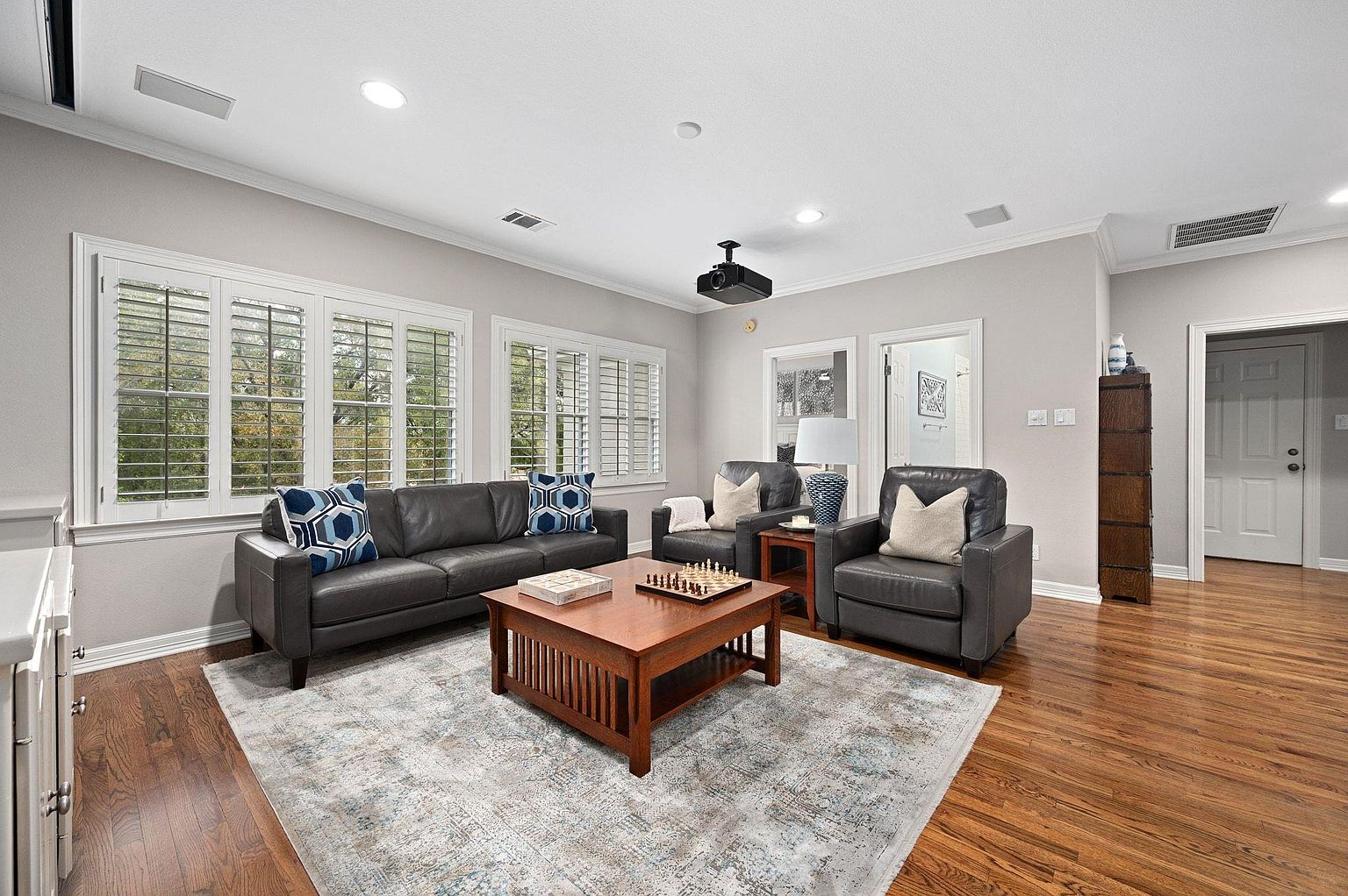 This is an interior shot of a living room featuring a dark leather sofa with patterned pillows, two matching armchairs, and a wooden coffee table with a chess set. A large area rug anchors the seating arrangement, and natural light streams in through shuttered windows. The room has a cozy and inviting atmosphere, perfect for relaxation and entertainment.