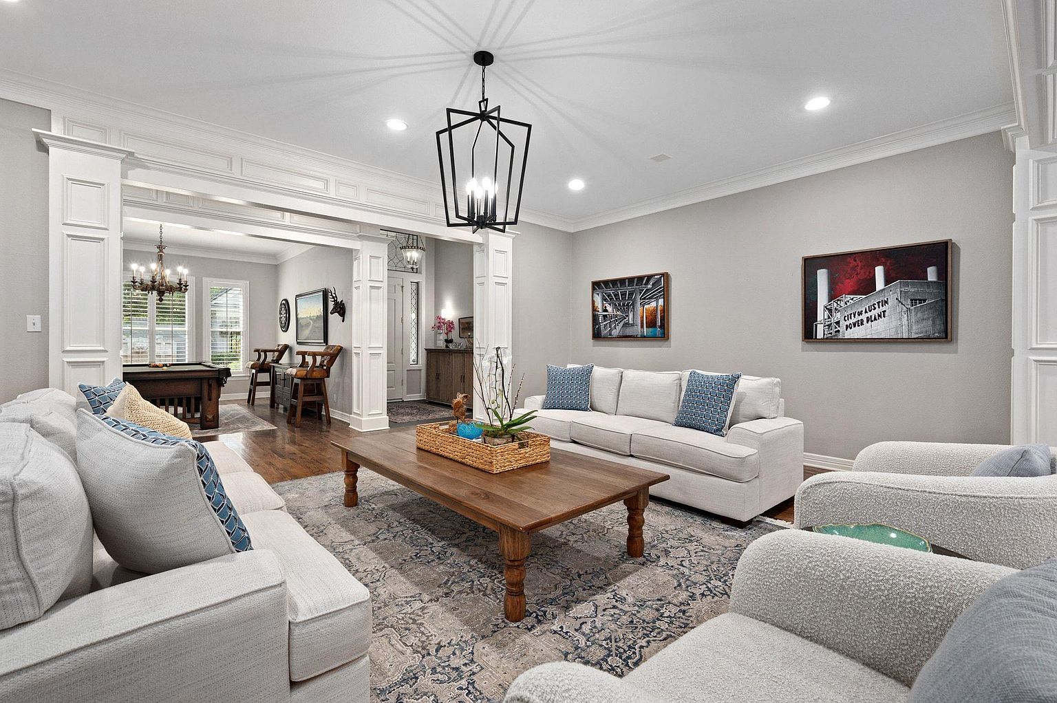 This is an interior shot of a living room, showcasing a spacious and well-lit area with a blend of traditional and contemporary elements. The room features comfortable seating arrangements, including sofas and armchairs, arranged around a wooden coffee table on an area rug. Architectural details like the white columns and trim add to the room's elegance, while artwork on the walls and a modern chandelier contribute to the overall aesthetic.