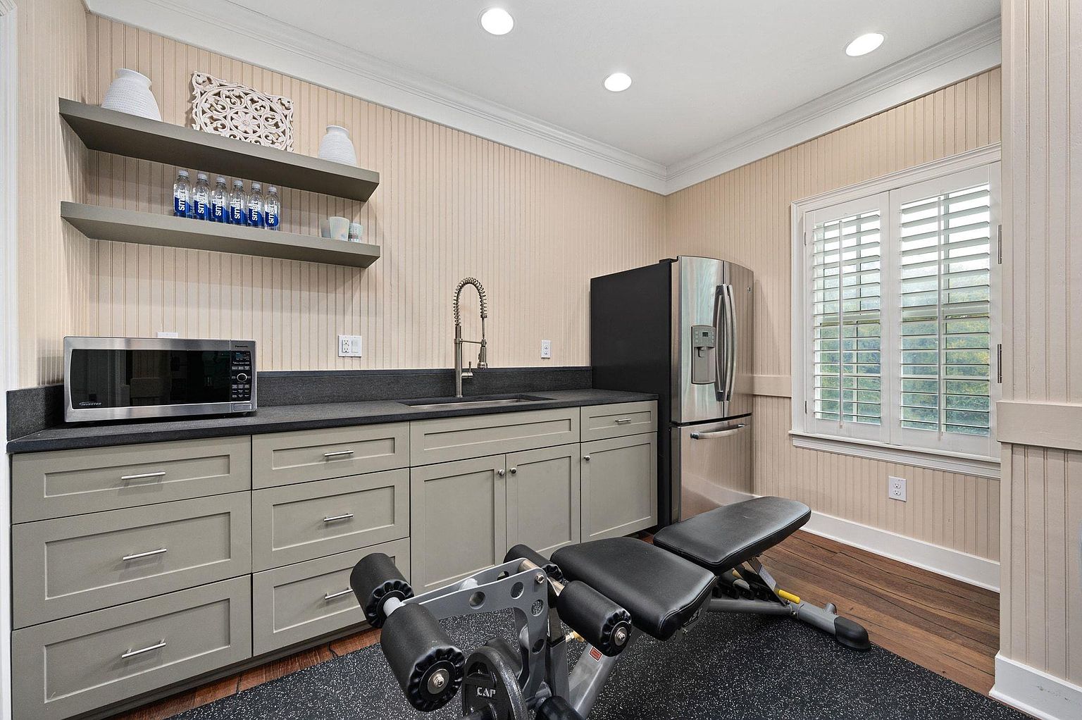 This interior shot showcases a well-equipped home gym area. The space features a wet bar with gray cabinetry, a dark countertop, a stainless steel refrigerator, and a microwave. A weight bench sits on a black rubber floor, and a window with white shutters provides natural light, creating a functional and inviting workout space.