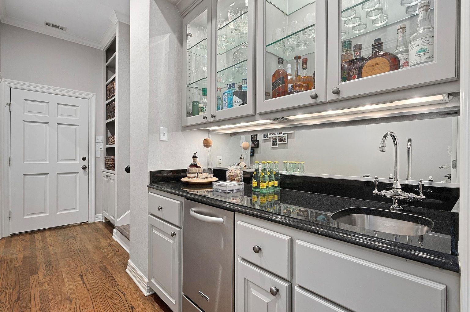 This interior shot showcases a stylish bar area with white cabinetry, a black countertop, and glass-front cabinets displaying glassware and liquor bottles. A stainless steel sink and faucet are visible, along with decorative items and beverage bottles on the counter. The overall impression is one of sophistication and functionality, perfect for entertaining.