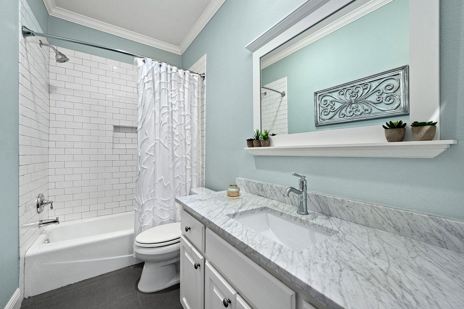 This is a well-lit bathroom featuring a white vanity with a marble countertop and a rectangular sink. A large mirror hangs above the vanity, reflecting the shower/tub area with white subway tiles and a decorative shower curtain. The walls are painted a light blue, creating a clean and refreshing atmosphere.