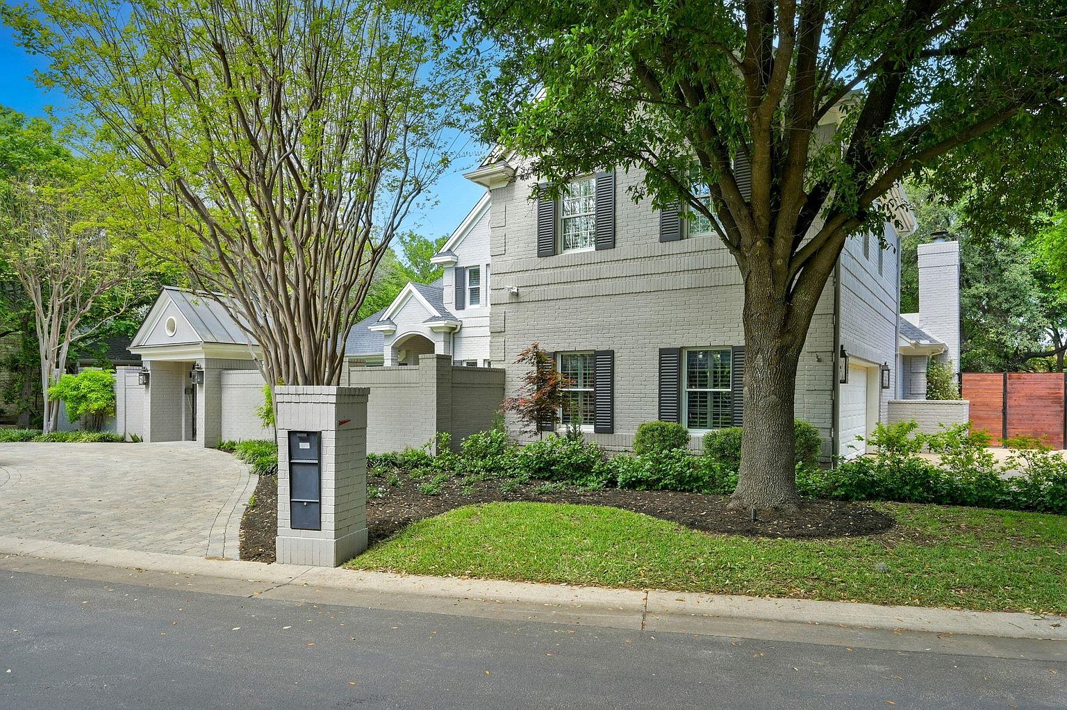This is a front exterior view of a two-story brick house with black shutters and a well-manicured lawn. A brick mailbox stands at the edge of the property, and mature trees frame the house, adding to its curb appeal. The driveway is paved with stone and leads to a garage.