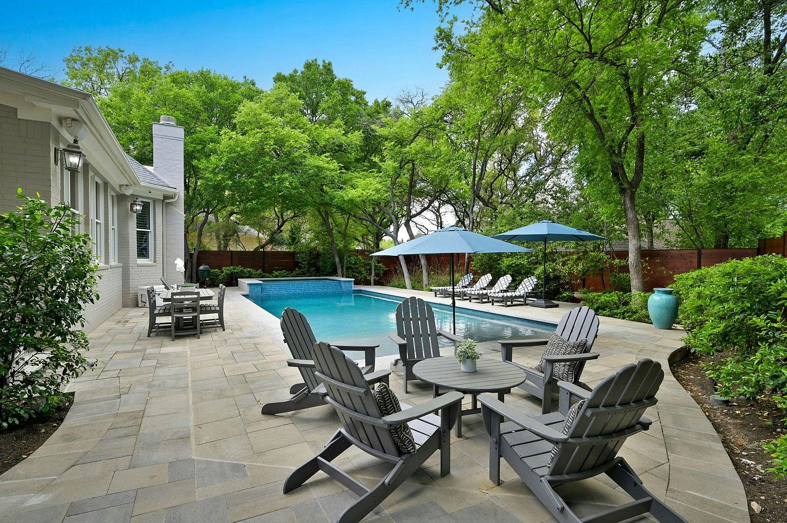 This image showcases a luxurious backyard pool area, complete with a patio furnished with comfortable seating around a small table. The pool is surrounded by lush greenery and mature trees, providing a private and serene atmosphere. Lounge chairs with umbrellas are placed near the pool, suggesting relaxation and leisure.