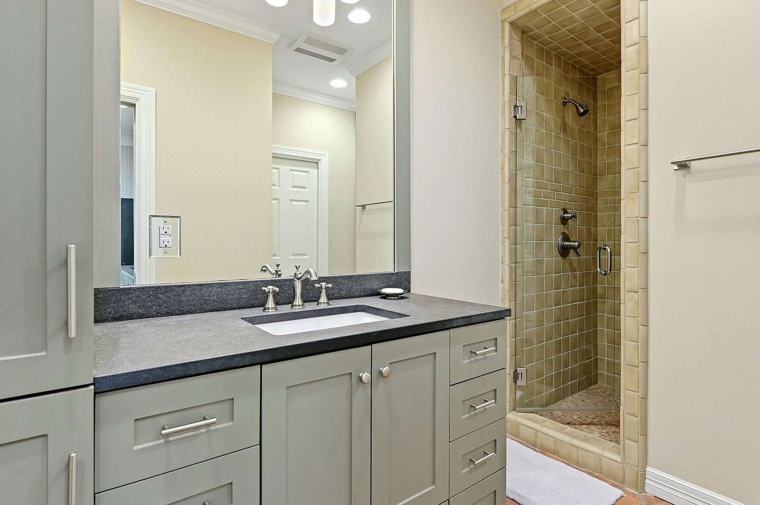 This is a well-lit primary bathroom featuring a gray vanity with a dark countertop and a white undermount sink. A large mirror reflects the vanity area and a glimpse of a nearby door. To the right, a tiled shower with a glass door adds a touch of luxury to the space.