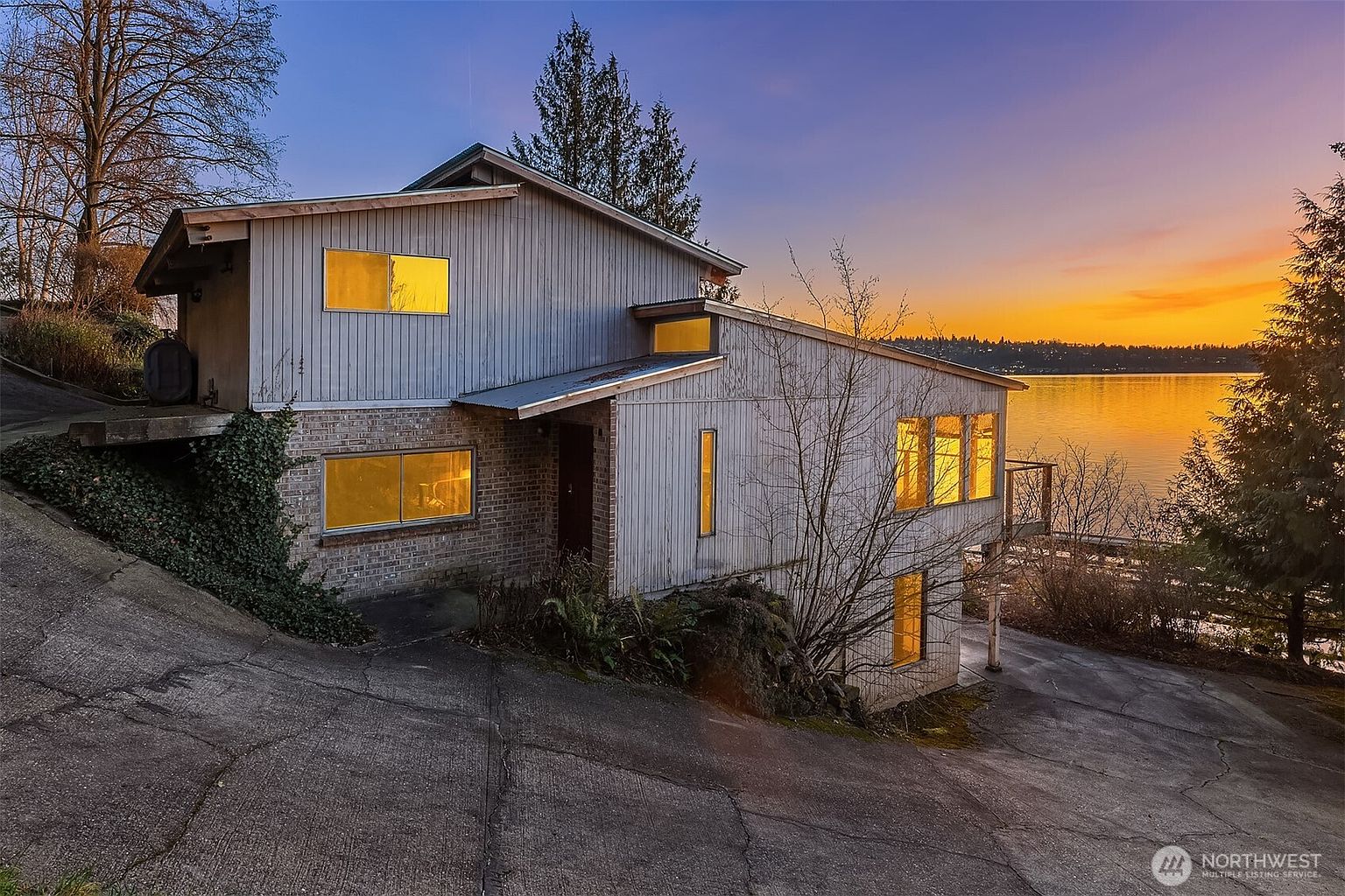 This striking multi-level home features a unique contemporary architectural design with a mix of brick and vertical wood siding, set against a dramatic sunset over the water. The perspective captures the home from the driveway, highlighting its tiered structure, large windows glowing with warm interior light, and its prime waterfront location. The overall impression is one of modern elegance integrated into a serene, natural landscape.