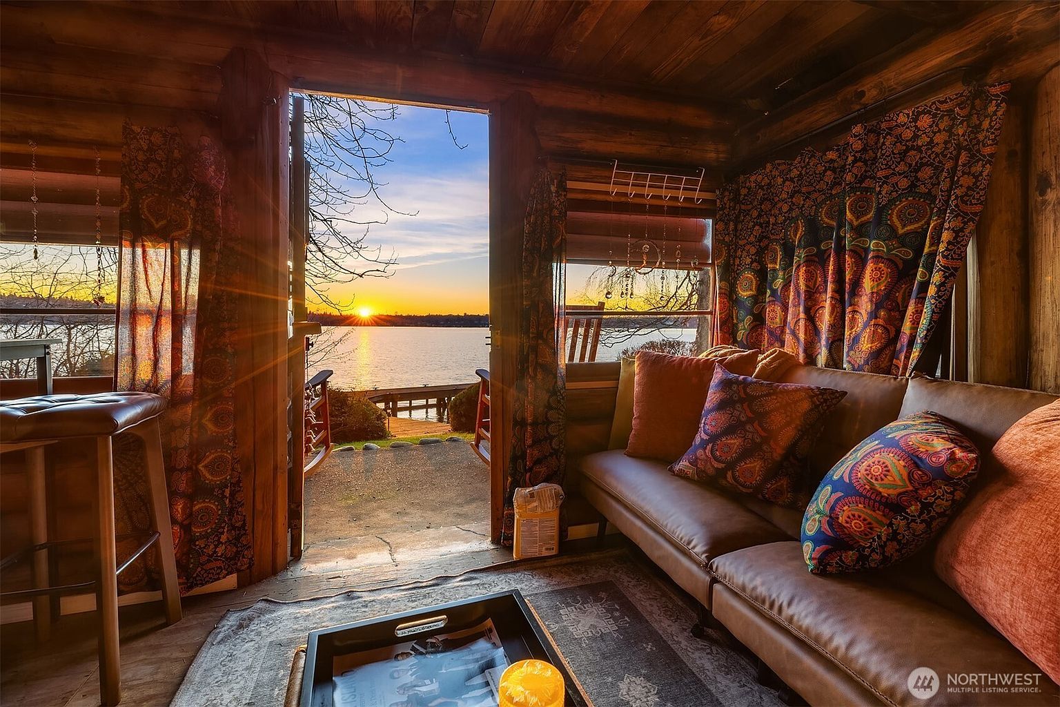 This cozy, rustic living space features warm log walls and a comfortable leather sofa adorned with vibrant, patterned throw pillows. The room opens directly onto a scenic waterfront view through a wide doorway, capturing a breathtaking sunset over the lake. The combination of bohemian textiles and natural wood creates an inviting, tranquil atmosphere perfect for relaxation.