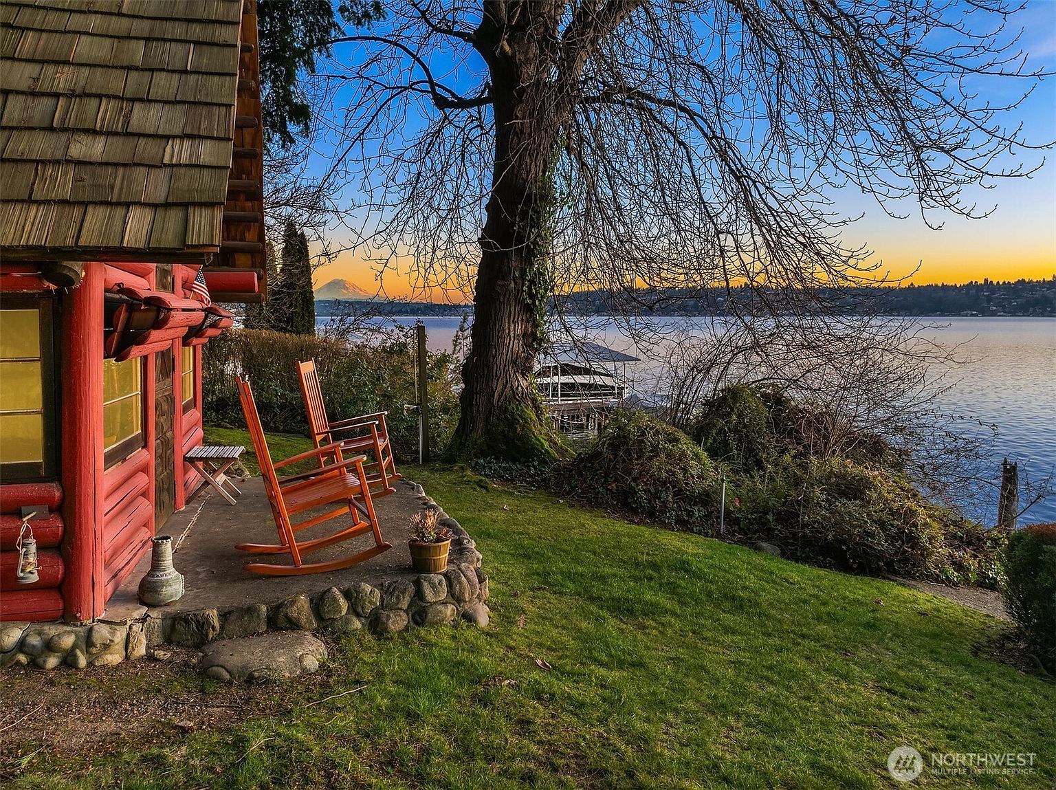 This picturesque scene captures a rustic log cabin porch overlooking a serene lake at sunset, with the iconic silhouette of Mount Rainier visible in the distance. The foreground features two wooden rocking chairs resting on a stone-bordered patio, set against a lush green lawn and a large, mature tree. The warm, golden hour lighting creates a tranquil and inviting atmosphere, highlighting the home's charming waterfront location.
