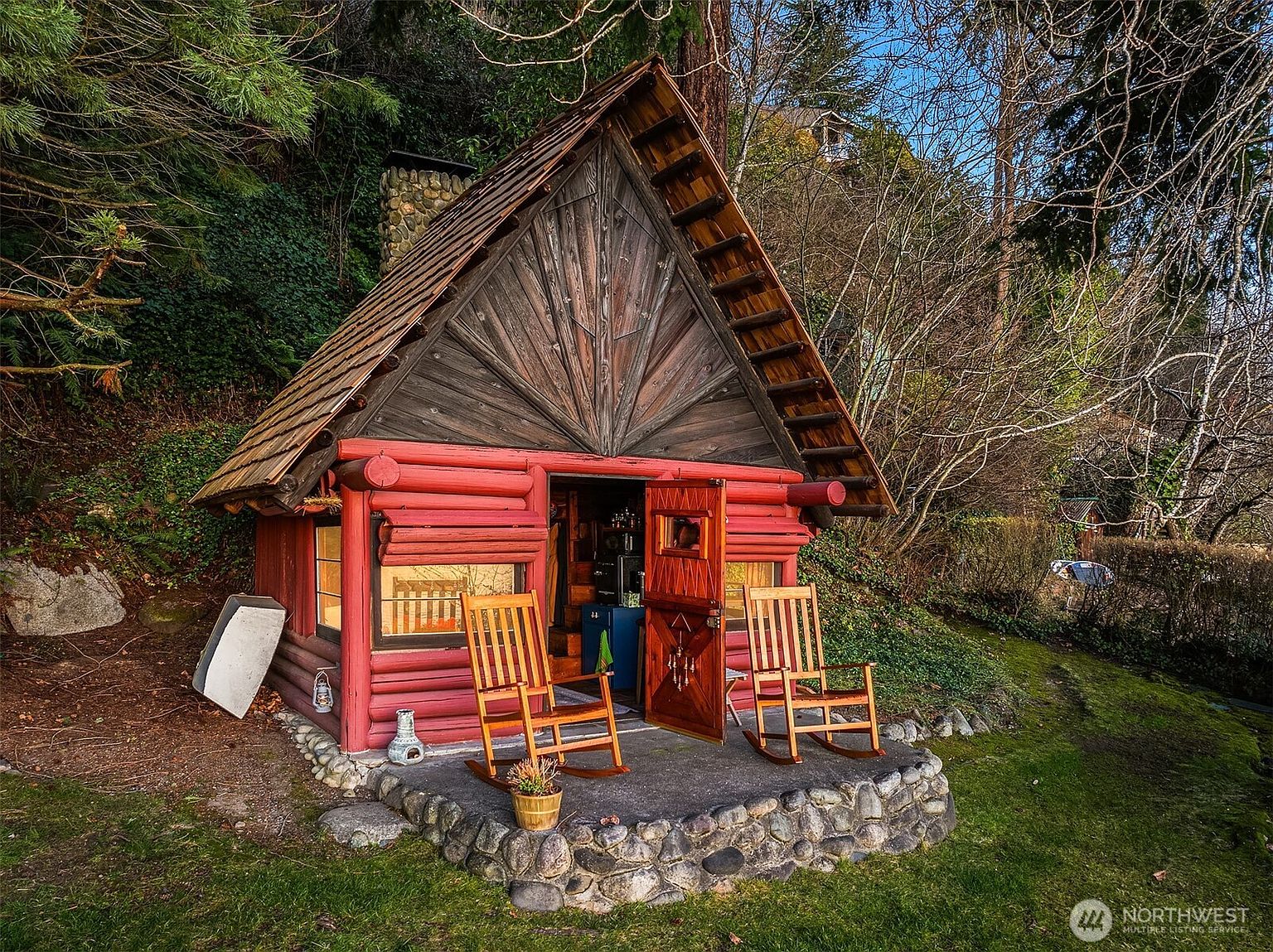 This charming, rustic A-frame cabin features vibrant red log walls and a unique sunburst-patterned wooden gable. A stone-bordered patio sits in front, furnished with two wooden rocking chairs, creating a cozy and inviting outdoor space nestled against a wooded hillside. The scene captures a peaceful, storybook aesthetic perfect for a secluded retreat.
