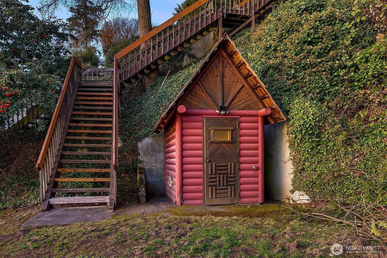 This unique outdoor space features a rustic, red-painted log cabin-style shed nestled against a hillside covered in lush ivy. A wooden staircase ascends the steep slope beside the shed, creating a charming, storybook-like aesthetic. The scene is captured from a ground-level perspective, highlighting the contrast between the natural greenery and the handcrafted architectural structure.