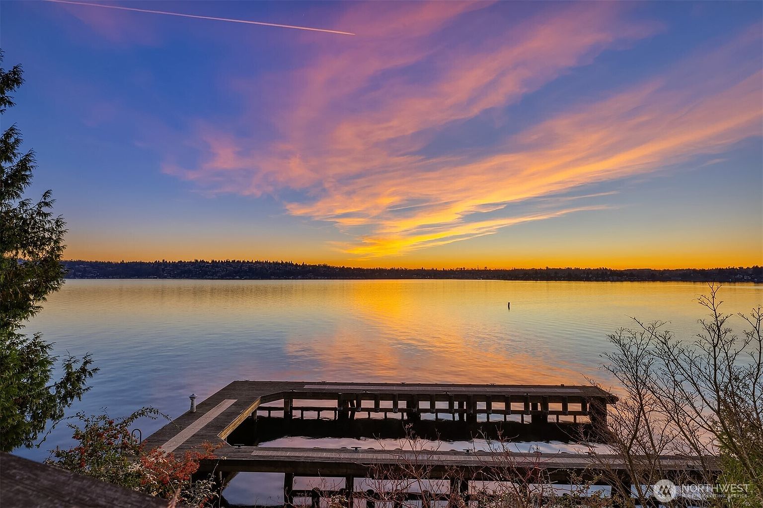 This scenic shot captures a wooden dock extending into a calm, expansive lake during a vibrant sunset. The sky is painted with brilliant hues of orange, pink, and deep blue, reflecting beautifully on the water's surface. Framed by lush trees on the left and bare branches on the right, the perspective offers a serene and picturesque view from an elevated deck, highlighting the property's prime waterfront location.