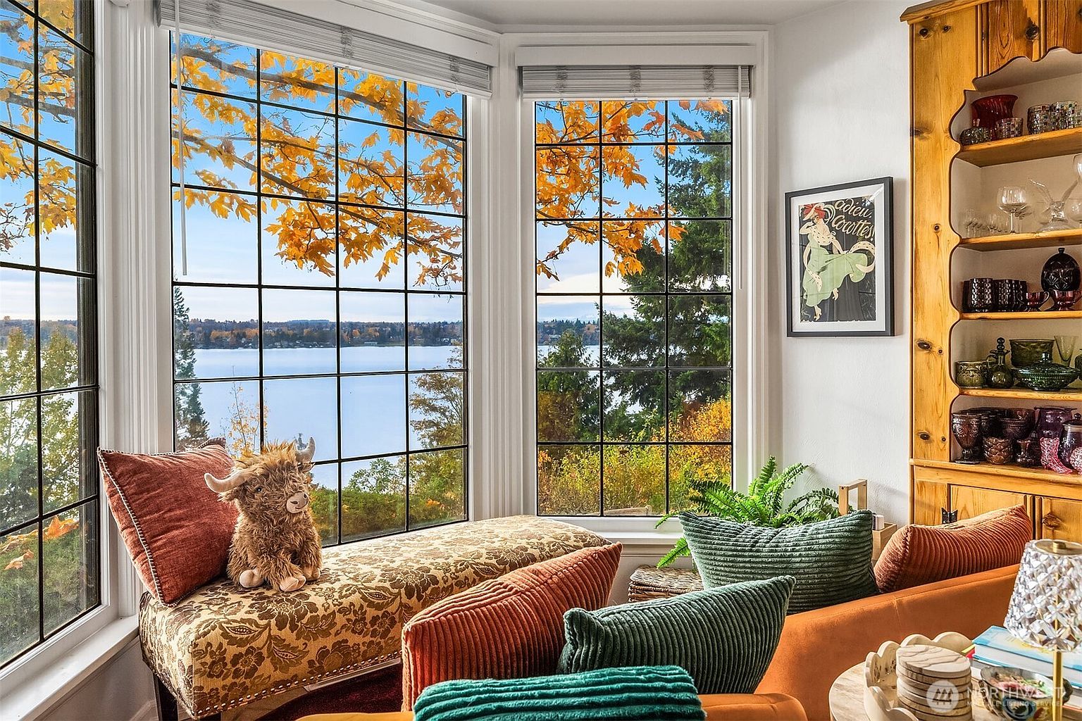 This cozy living room area features a large bay window that frames a stunning, panoramic view of a lake surrounded by autumn foliage. The space is furnished with a patterned window seat adorned with a plush highland cow toy, complemented by warm-toned velvet pillows and a wooden built-in shelving unit displaying decorative glassware. The overall atmosphere is inviting and serene, perfectly blending comfortable indoor living with the natural beauty of the outdoors.