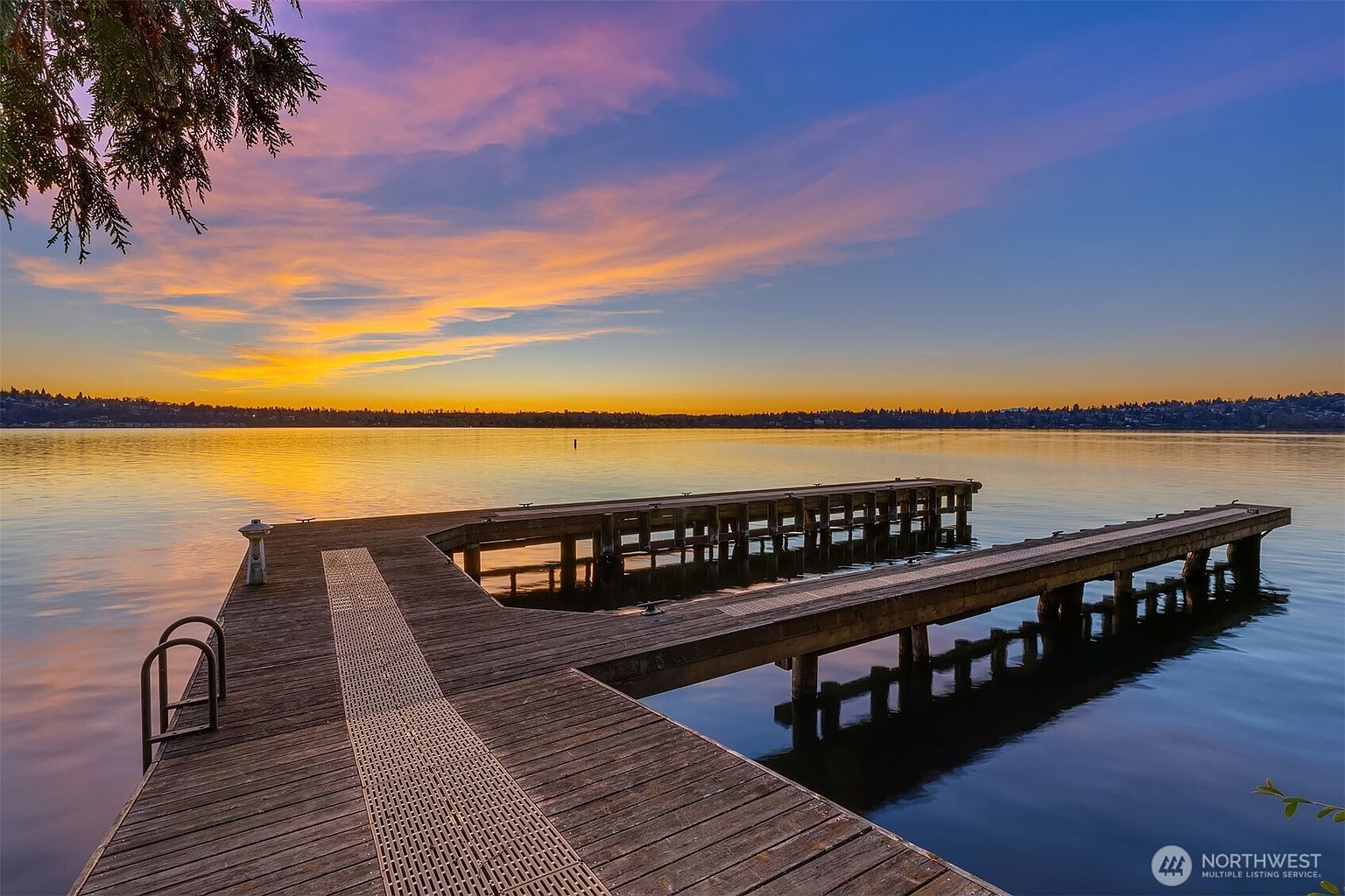 This image captures a serene, wooden boat dock extending into a calm lake during a vibrant sunset. The perspective is from the dock looking out toward the horizon, where the sky is painted in hues of orange, pink, and deep blue, reflecting beautifully on the water's surface. The scene evokes a peaceful, luxurious waterfront lifestyle, highlighting the property's direct access to the lake.