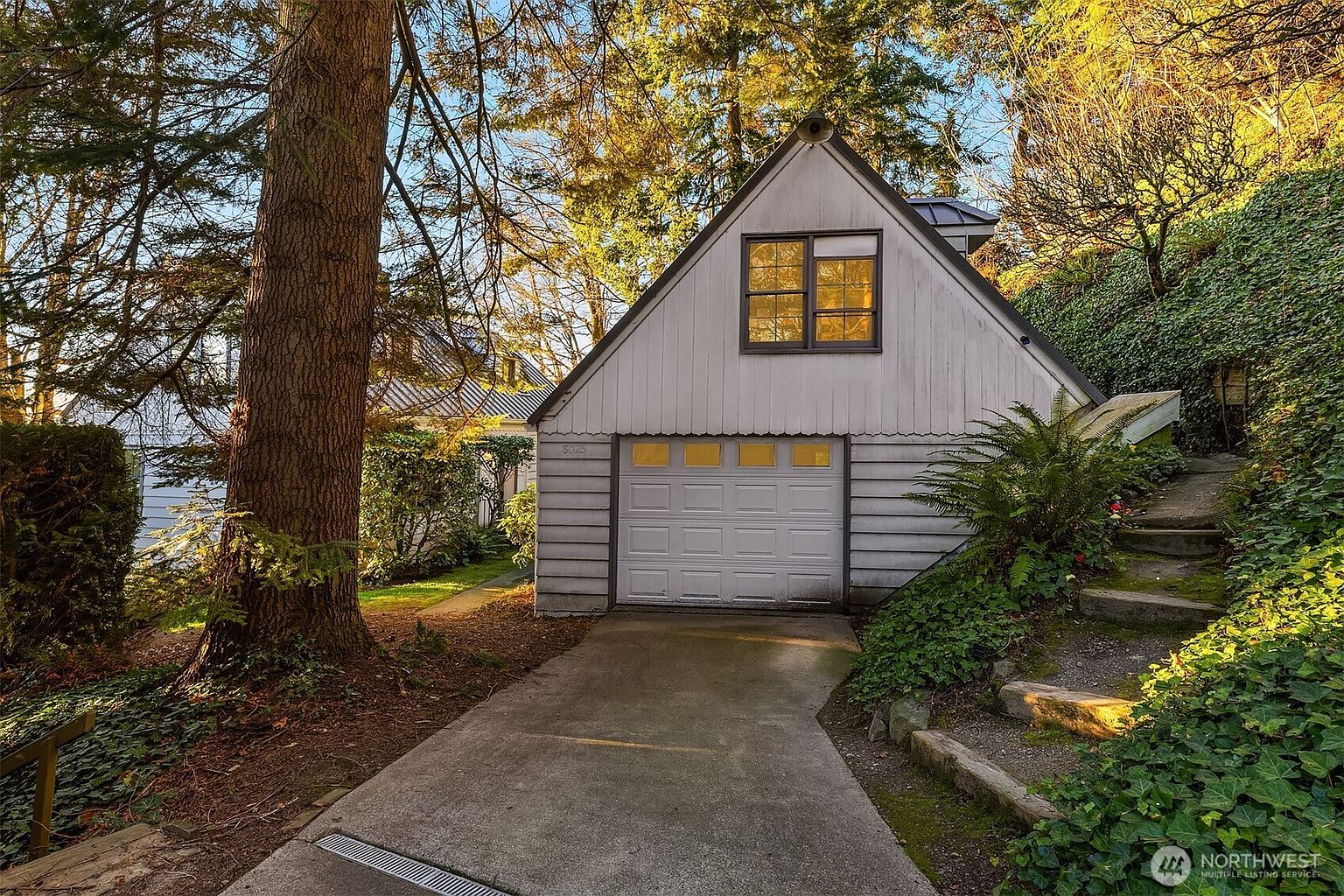 This image captures a detached, A-frame style garage nestled into a lush, wooded hillside. The structure features light-colored vertical siding, a single-car garage door, and an upper-level window, all set against a backdrop of mature trees and dense ivy-covered slopes. A concrete driveway leads up to the garage, while stone steps on the right provide access to the elevated landscape, creating a secluded and rustic atmosphere.