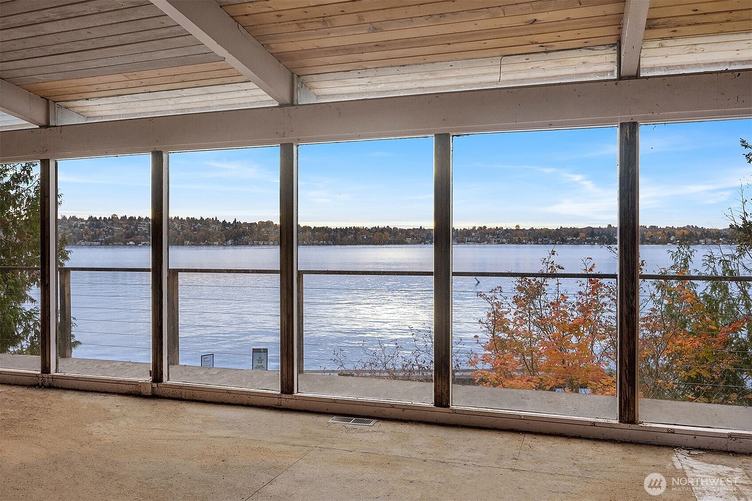This sunroom features a wall of floor-to-ceiling windows that provide a panoramic, unobstructed view of a serene lake and the distant shoreline. The space has a rustic, unfinished aesthetic with exposed wooden ceiling beams and a simple concrete floor, emphasizing the natural beauty of the waterfront setting. The perspective is from inside looking out, highlighting the seamless connection between the interior living space and the tranquil outdoor environment.