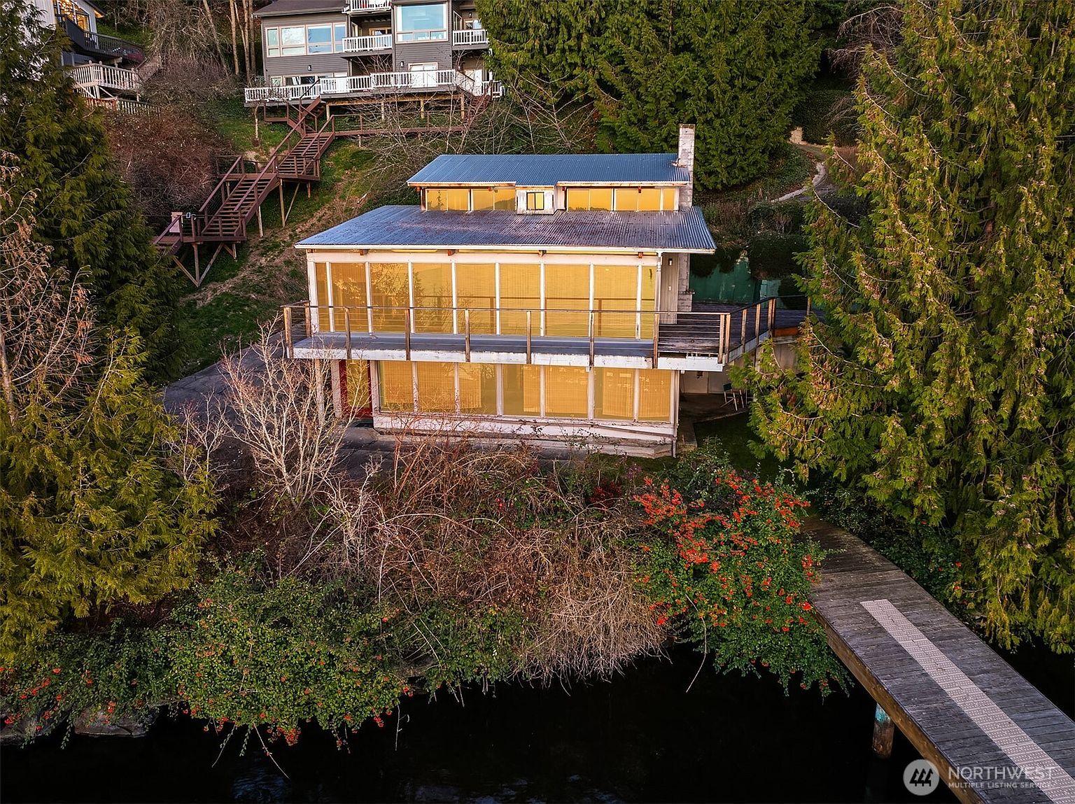 This aerial perspective captures a multi-level waterfront home nestled into a lush, wooded hillside. The house features a prominent deck overlooking the water, large floor-to-ceiling windows with a warm, golden reflection, and a private wooden dock extending into the lake. The setting is serene and secluded, emphasizing the property's direct access to the water and its integration with the surrounding natural landscape.