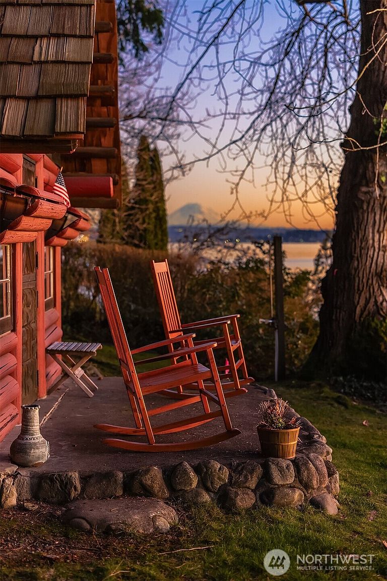 This inviting porch area features two classic wooden rocking chairs set against a rustic log cabin exterior, complete with a small American flag. The stone-bordered patio overlooks a serene waterfront view at sunset, with a distant mountain peak visible on the horizon. The scene is framed by a large tree and soft, warm lighting, creating a peaceful and picturesque atmosphere perfect for relaxation.