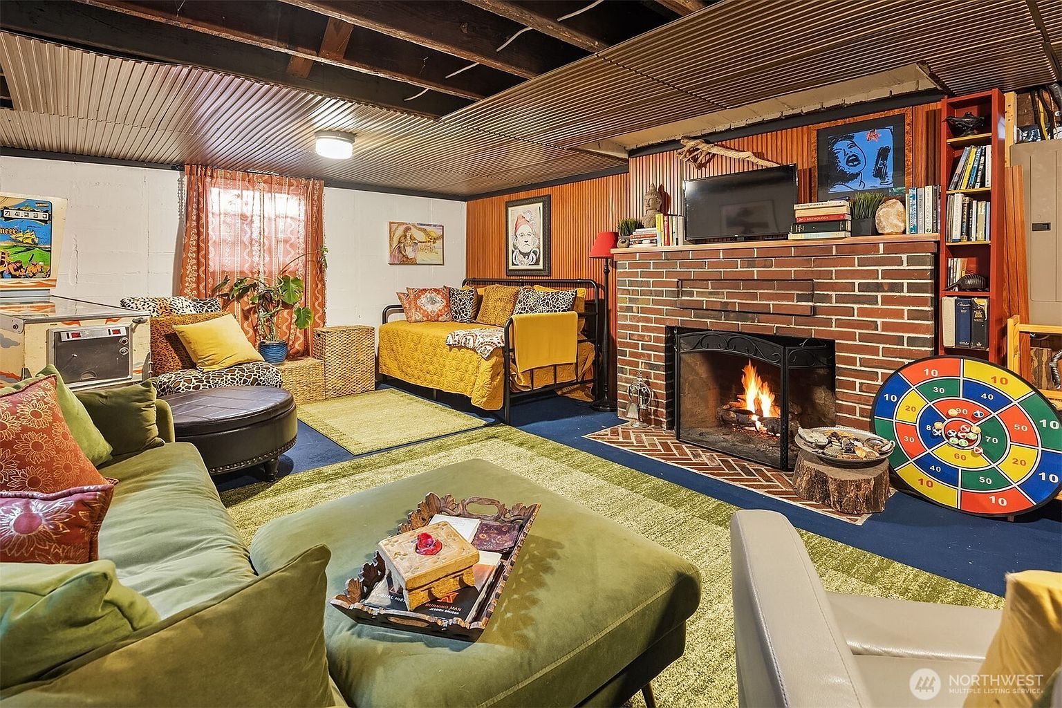 This cozy, eclectic basement living area features a warm brick fireplace as its focal point, complemented by a vibrant green velvet sofa and a daybed with patterned textiles. The space is finished with corrugated metal ceilings and wood-paneled walls, creating a unique, retro-inspired lounge atmosphere complete with a pinball machine and a dartboard. The perspective is a wide-angle shot from the foreground, capturing the inviting and multi-functional layout of the room.