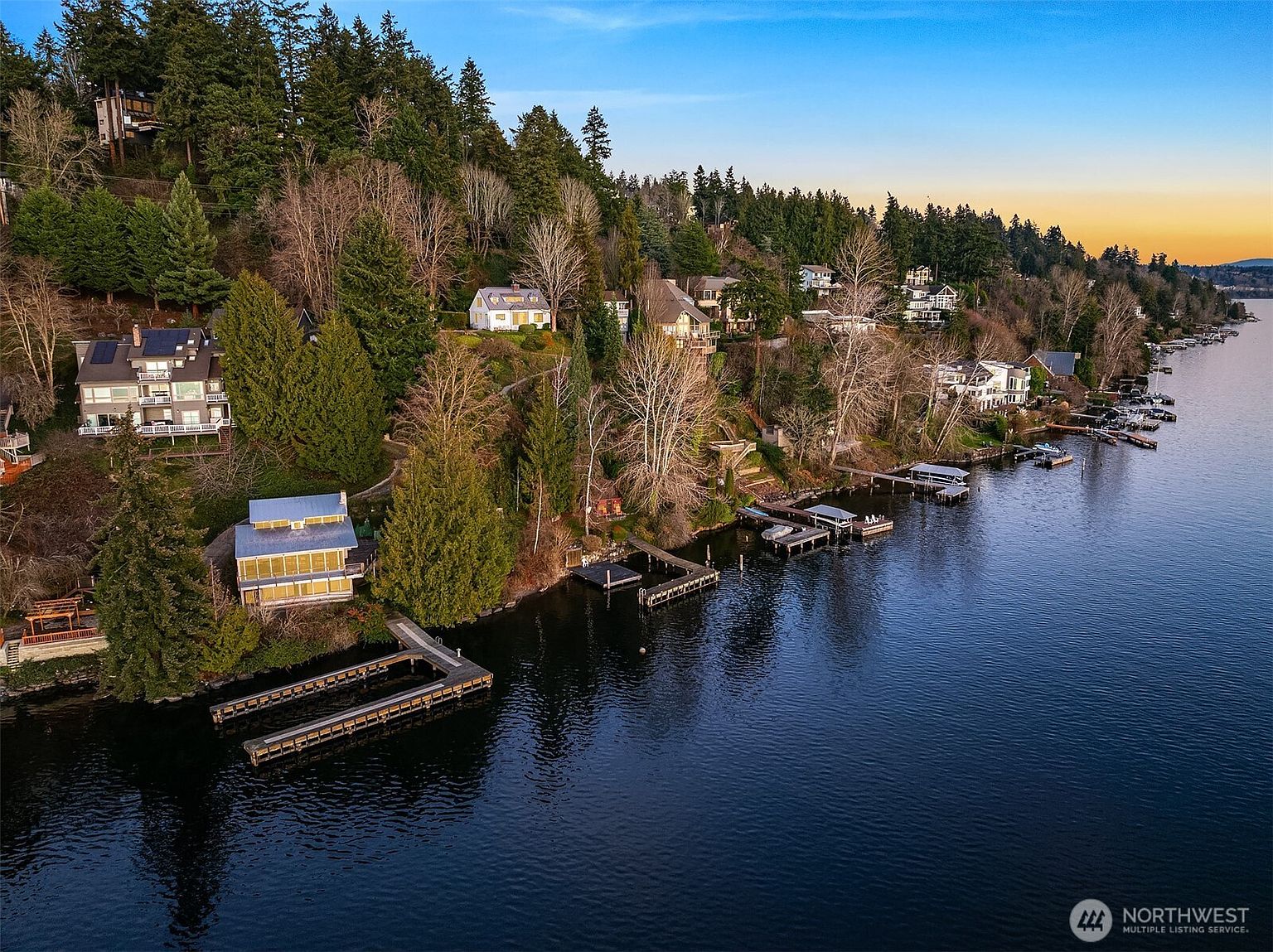 This high-angle aerial view captures a serene waterfront property nestled along a lush, tree-covered hillside. The scene features a multi-level home with a private dock extending into the calm, dark water, surrounded by a landscape of mature evergreens and neighboring residences. The golden hour lighting casts a warm glow over the shoreline, emphasizing the peaceful and secluded nature of this lakeside setting.