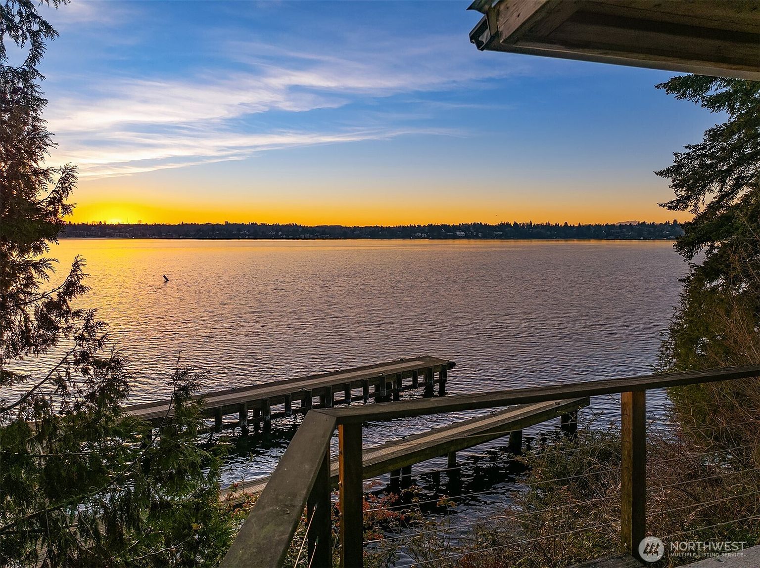 This image captures a serene, elevated view from a wooden deck overlooking a calm lake at sunset. The perspective highlights a private dock extending into the water, framed by the natural silhouettes of trees and a vibrant, golden-hued horizon. The scene evokes a peaceful, high-end waterfront lifestyle, emphasizing the property's direct access to nature and scenic views.