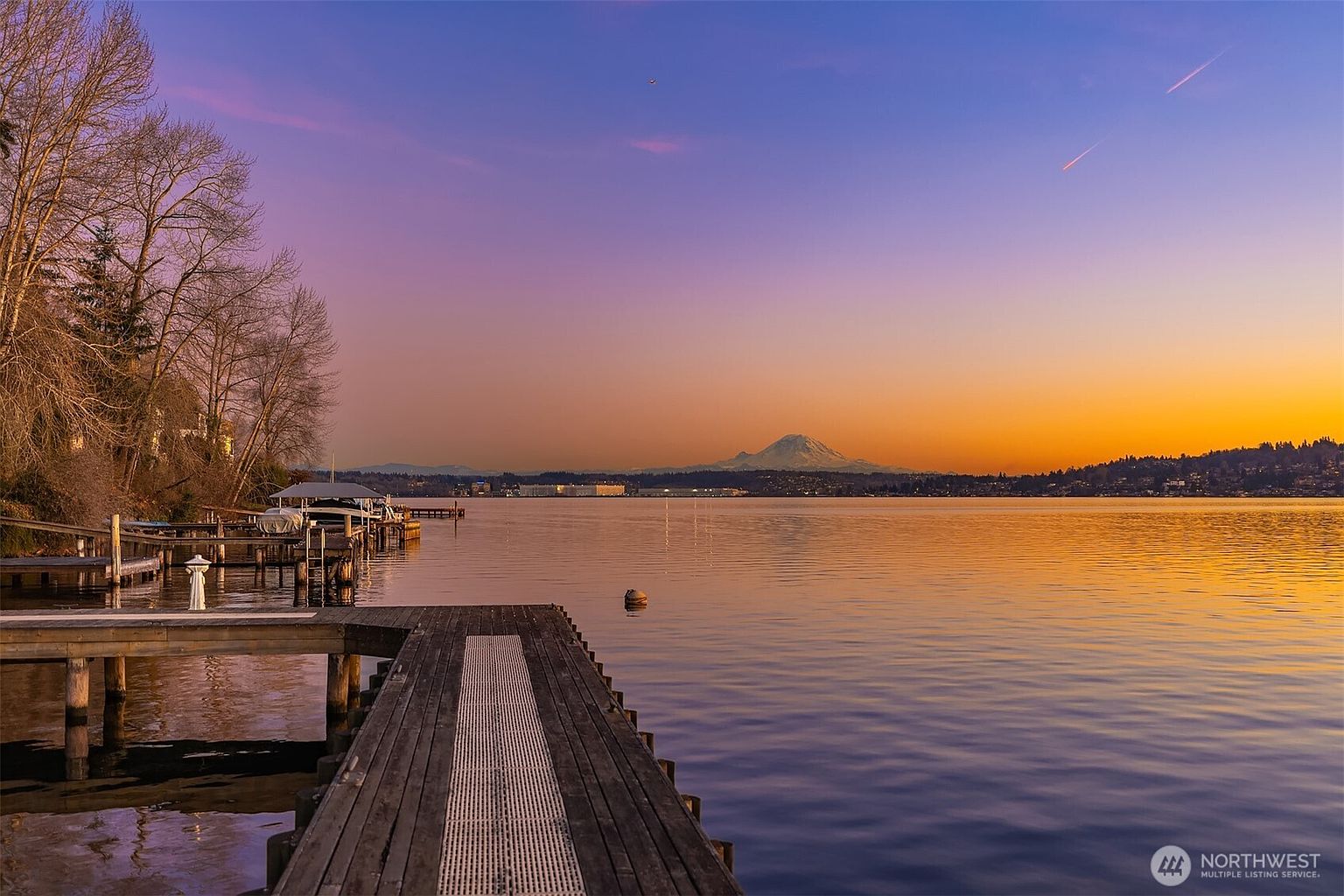 This stunning waterfront view captures a wooden dock extending into a calm lake at sunset, with the iconic silhouette of Mount Rainier visible in the distance. The scene features a serene, golden-hour atmosphere with soft purple and orange hues reflecting off the water's surface. The perspective is from the end of the dock, emphasizing the peaceful connection between the property and the natural landscape.