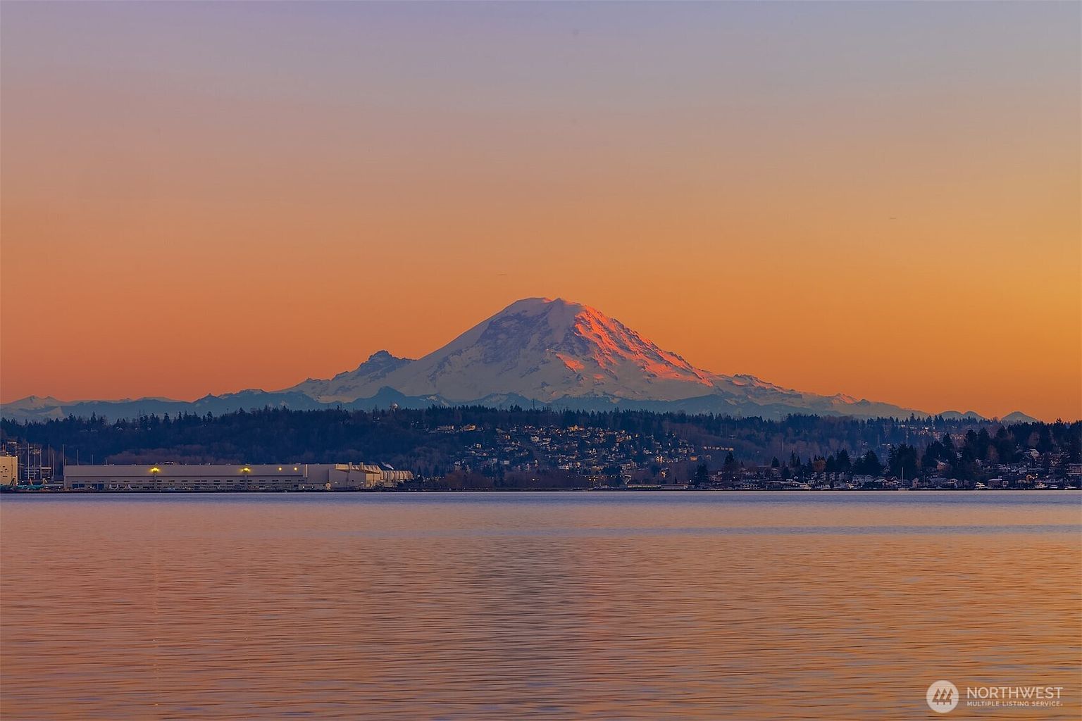 This scenic landscape shot captures a breathtaking view of a snow-capped mountain at sunset, reflecting a warm, golden glow across the calm waters of a bay. In the foreground, a tranquil shoreline features a residential area with houses nestled among trees and a large industrial building near the water's edge. The perspective is a wide, cinematic long shot that emphasizes the serene and majestic natural surroundings of the property's location.