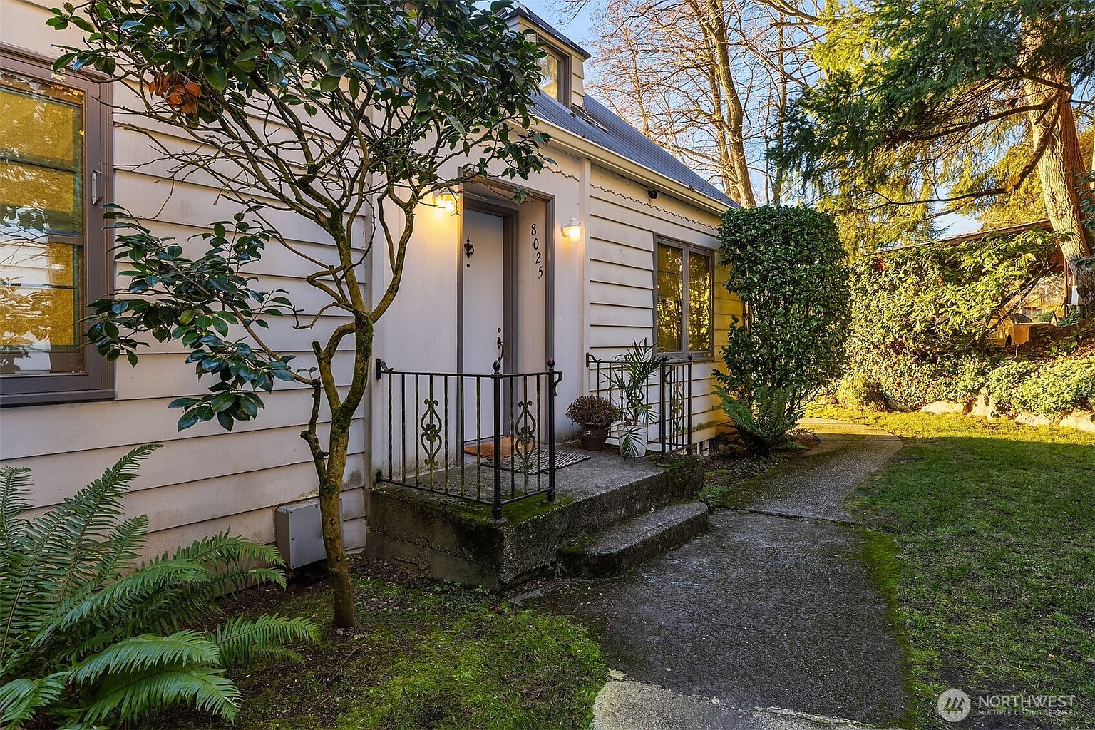 This charming entryway features a concrete porch with a classic black wrought-iron railing, leading to a white front door marked with the house number 8025. The exterior is finished with light-colored horizontal siding, complemented by a mature tree in the foreground and a lush, mossy garden path. The scene is captured from a slightly low angle, emphasizing the welcoming and well-maintained curb appeal of the home.