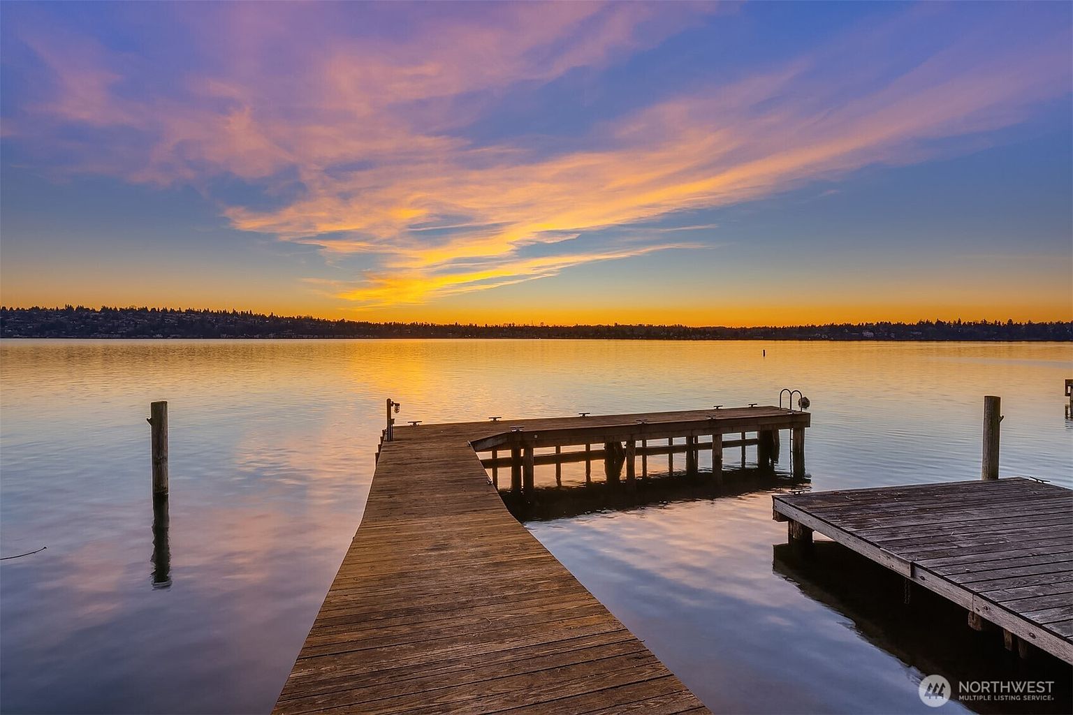 This serene waterfront view captures a wooden dock extending into a calm lake during a vibrant sunset. The sky is painted with hues of orange, purple, and blue, reflecting beautifully on the water's surface, creating a peaceful and picturesque atmosphere. The perspective is from the end of the dock, looking out toward the distant shoreline, emphasizing the tranquil lifestyle associated with waterfront property.
