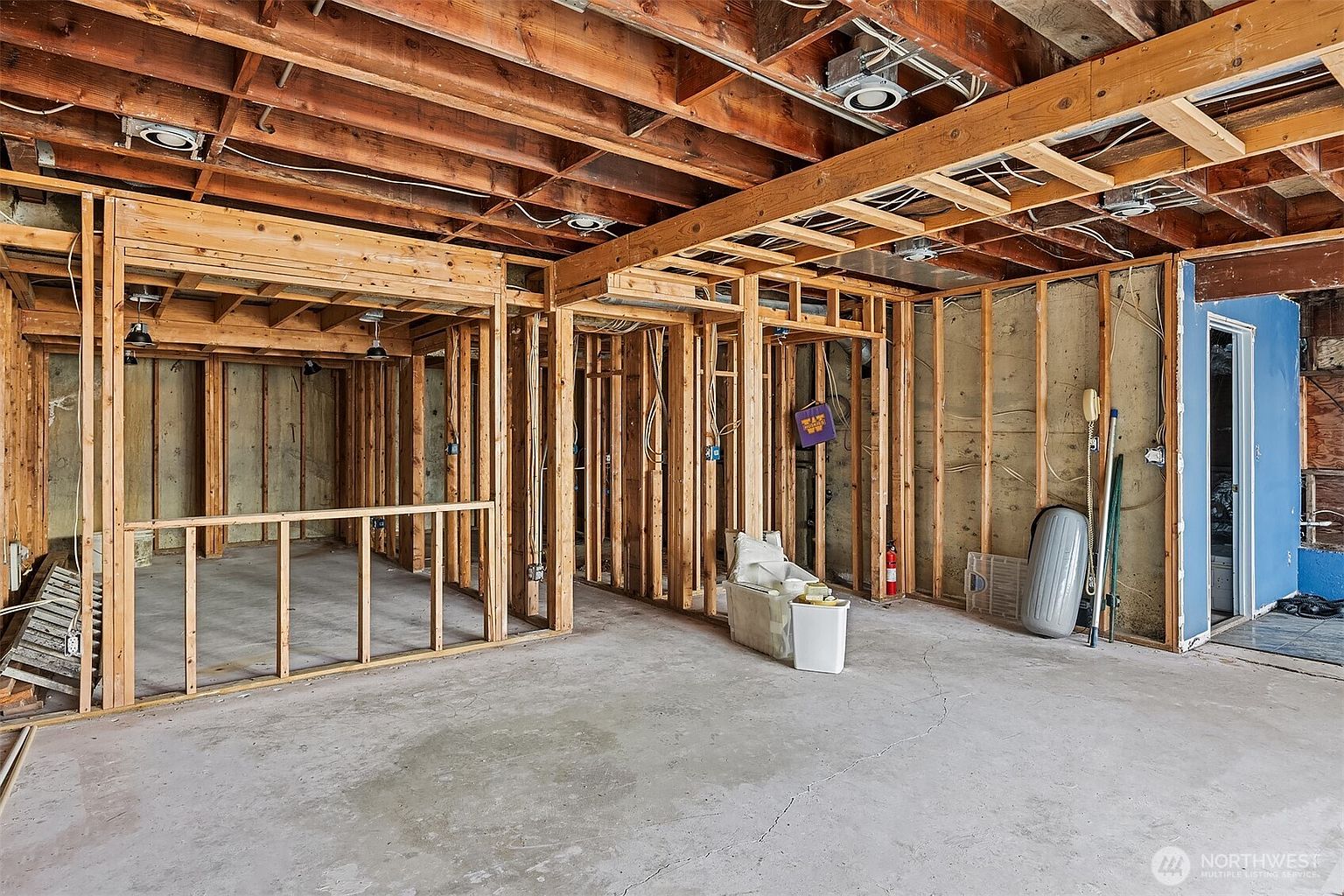 This image captures an unfinished basement space currently undergoing renovation, featuring exposed wooden framing, ceiling joists, and concrete flooring. The open-concept layout reveals the structural bones of the room, including electrical wiring and recessed lighting fixtures installed in the ceiling. The perspective is a wide-angle shot from the center of the room, highlighting the potential for a customizable living area.