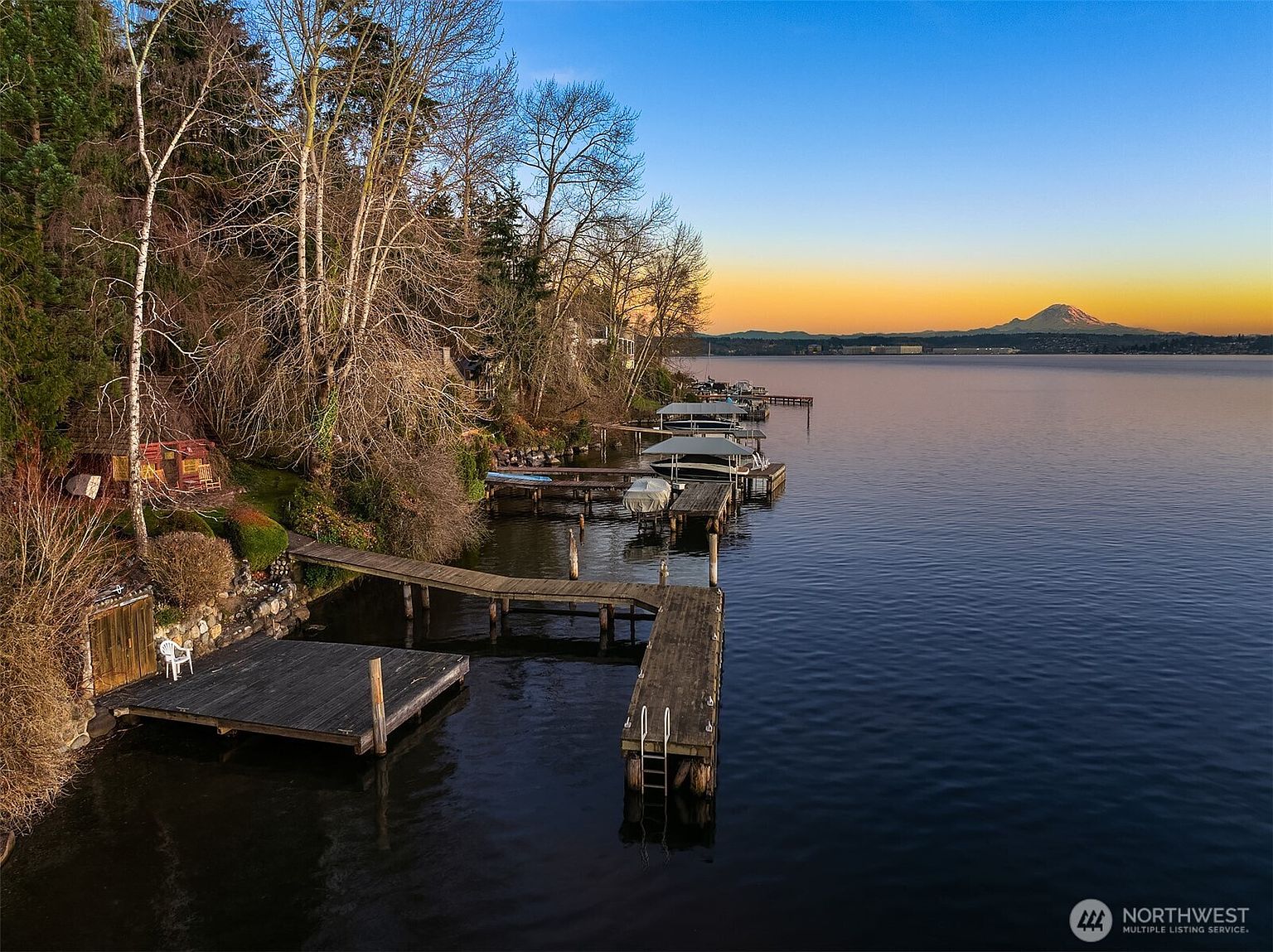 This aerial perspective captures a serene waterfront property featuring a private wooden dock extending into a calm lake at sunset. The scene highlights the lush, tree-lined shoreline and a distant, snow-capped mountain peak, creating a tranquil and picturesque atmosphere. The composition emphasizes the peaceful lifestyle and natural beauty associated with this prime lakeside location.