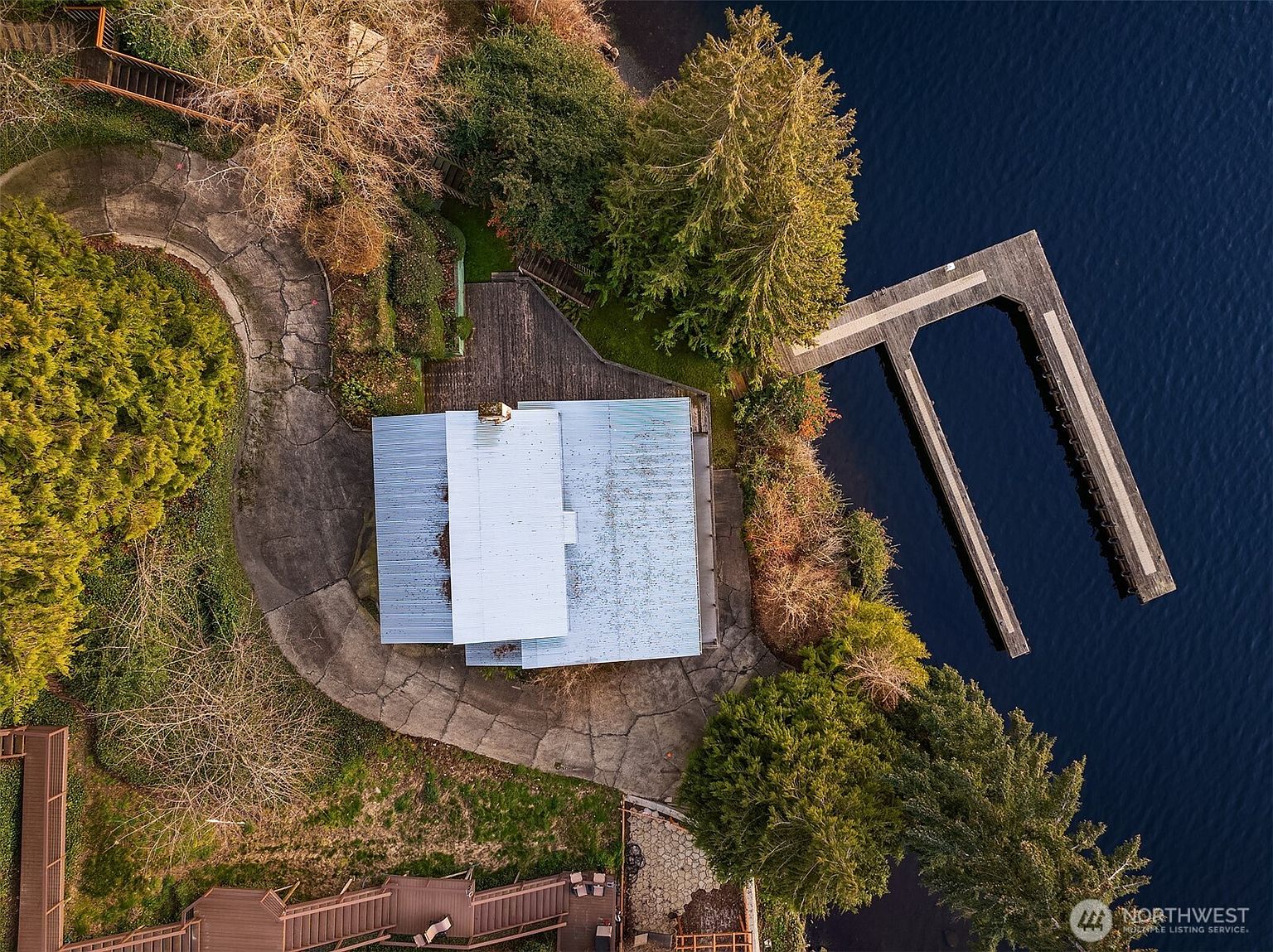 This high-angle aerial view captures a waterfront property featuring a house with a metallic roof nestled among mature trees and lush landscaping. A prominent U-shaped wooden dock extends directly into the dark, calm water, providing private access to the lake. The scene highlights the property's secluded, nature-focused setting and its direct connection to the shoreline.