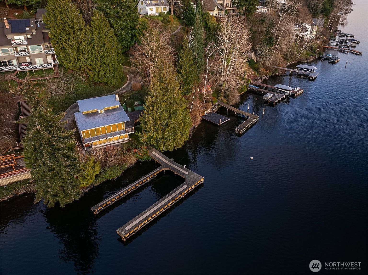 This high-angle aerial view captures a waterfront property featuring a distinctive multi-level home with a metal roof and expansive windows overlooking a private dock. The lush, wooded landscape slopes down to the water's edge, showcasing a serene and secluded setting. The perspective highlights the home's integration with the natural environment and its direct access to the lake.